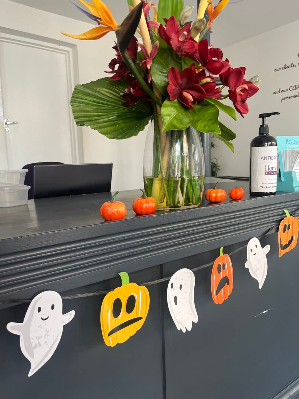 Halloween-themed decorations with ghost and pumpkin cutouts on a counter, vase with flowers, and hand sanitizer. - Home Instead
