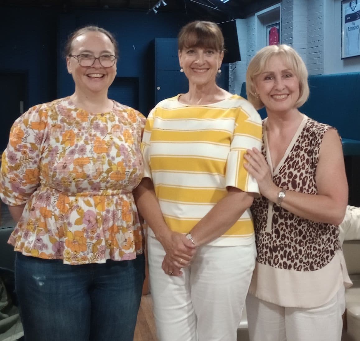 Three women smiling, standing together in a room with a blue wall and a mix of casual and patterned outfits. - Home Instead