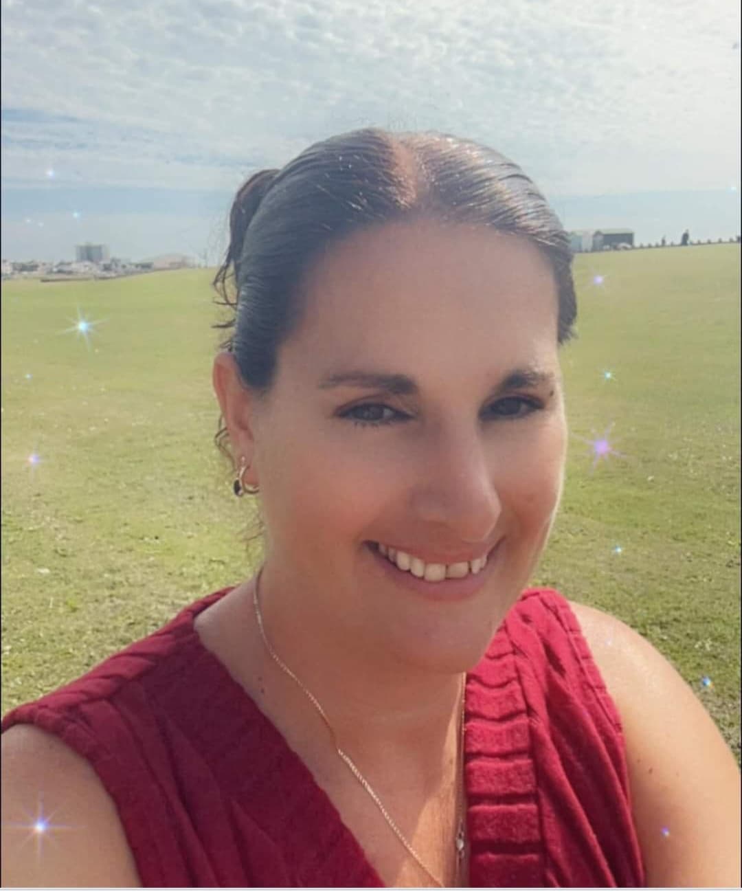 A woman in a red top smiles outdoors with a grassy field and blue sky in the background. - Home Instead