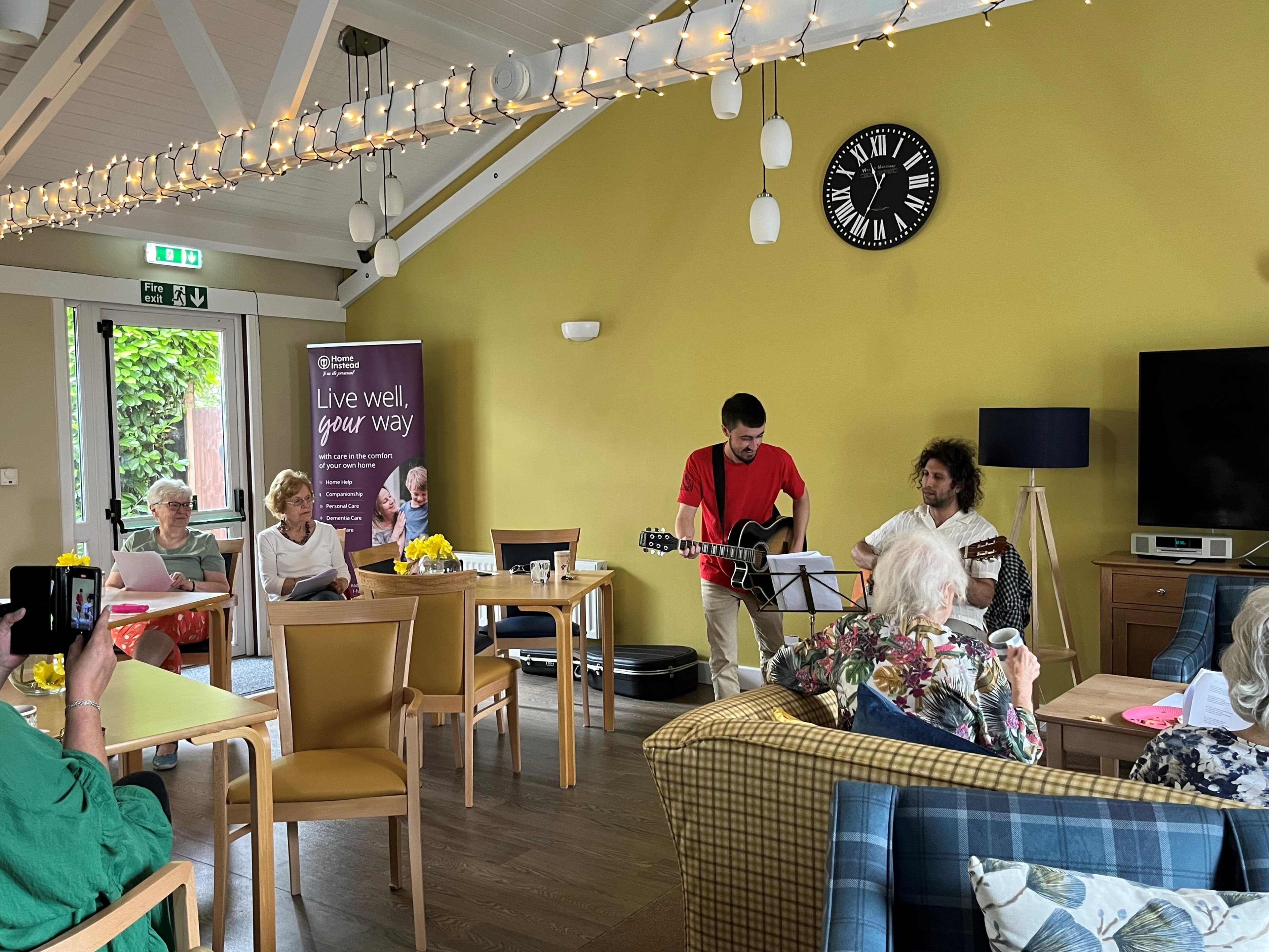 Musicians perform for seated seniors in a cozy, well-lit room with a clock and string lights. - Home Instead