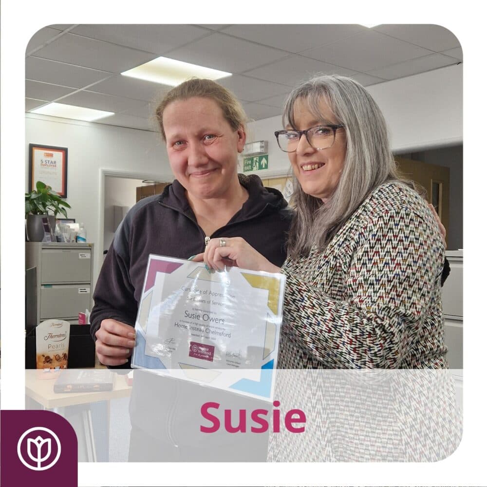 Two women smiling, one holding a certificate, standing in an office space with "Susie" written below. - Home Instead