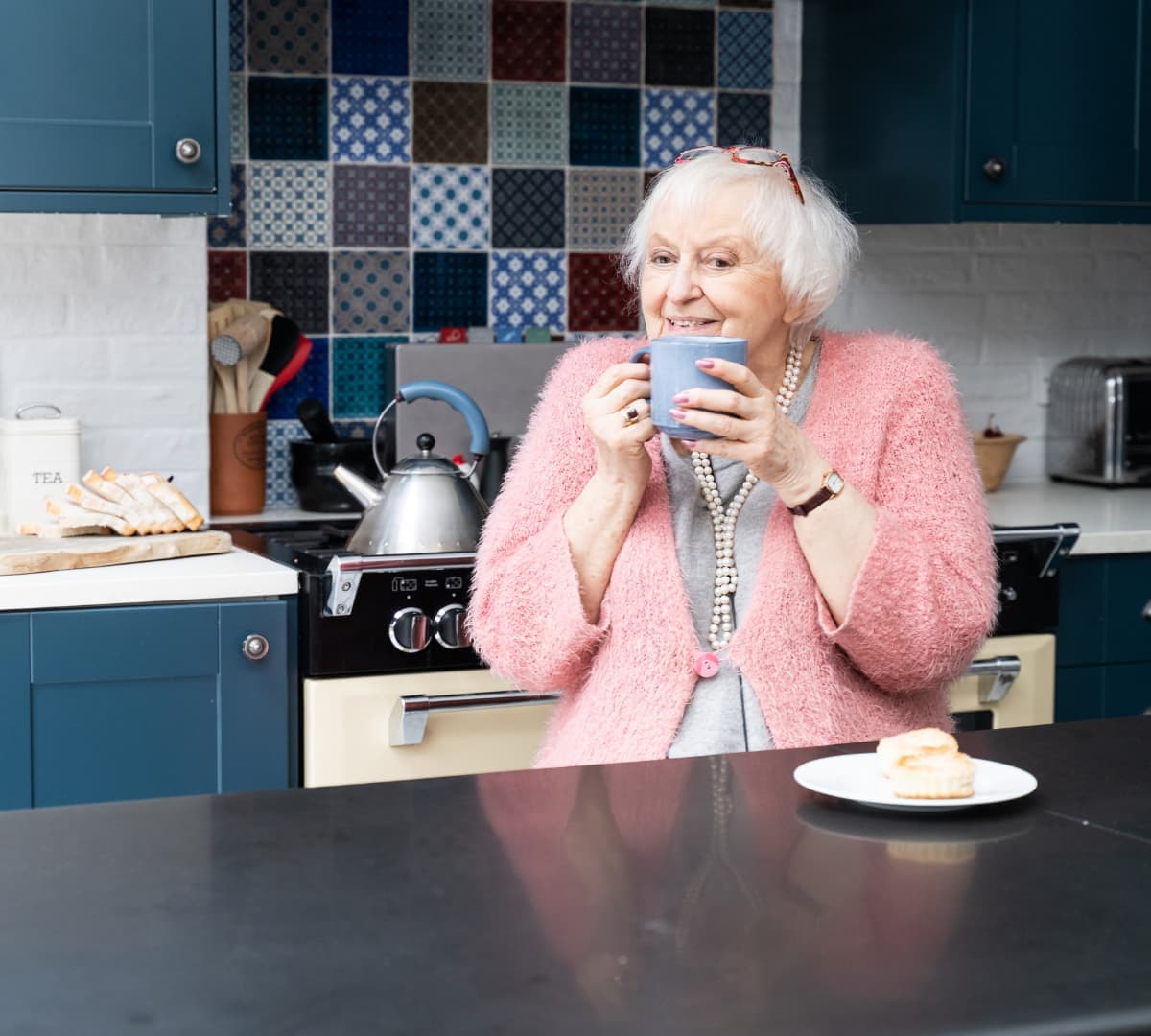A happy senior wearing pink sweater, smiling and sipping a cup coffee and eating scones in her kitchen