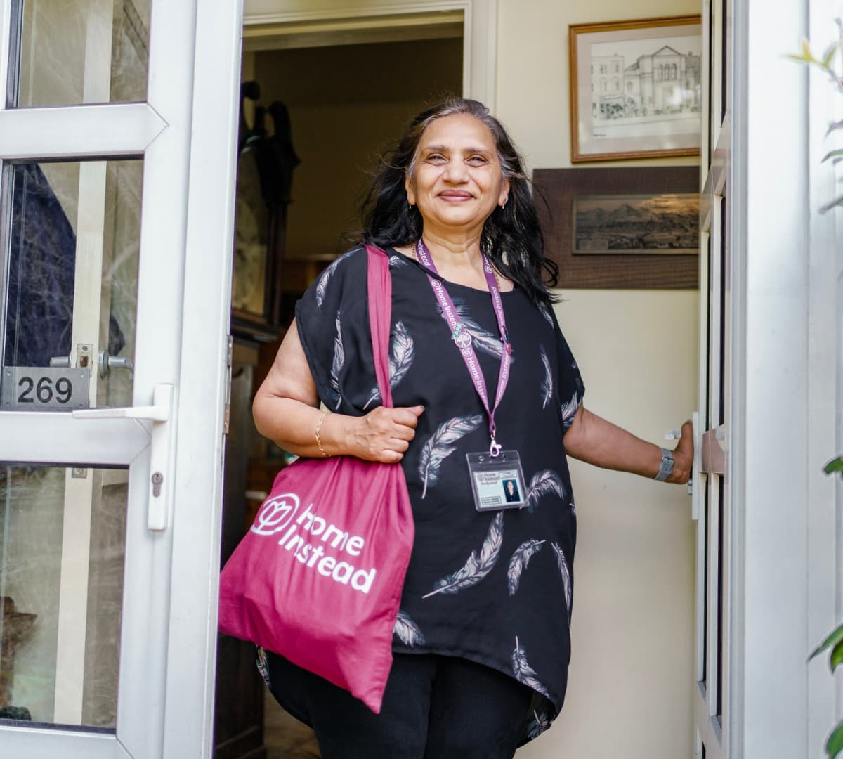 A smiling woman at the door with black long hair wearing black and carrying a Home Instead bag