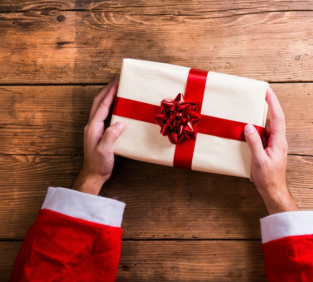 Two hands holding a christmas gift on a table in brown wrapper with red ribbon
