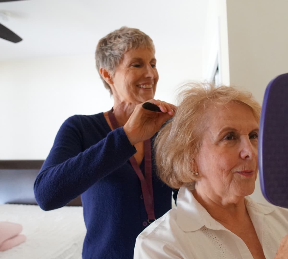 A carer with short hair wearing blue combing the hair of an older adult wearing white and holding a mirror
