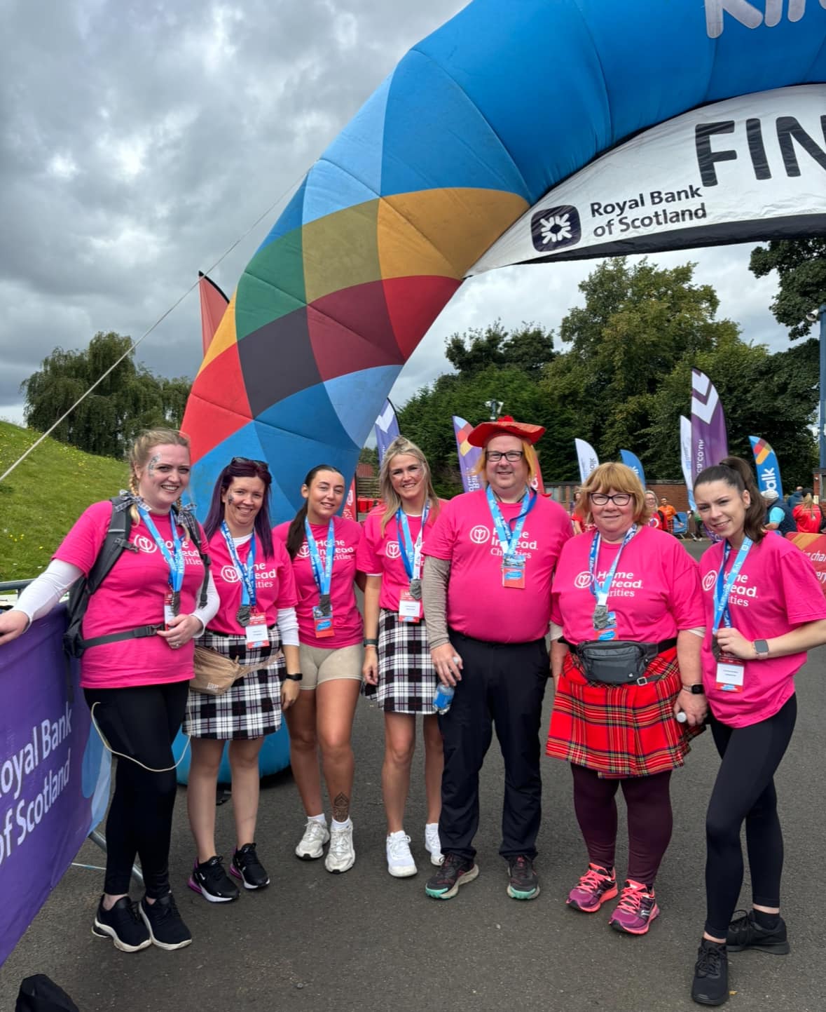 Group of people in pink shirts and kilts standing under a colorful finish line archway. - Home Instead