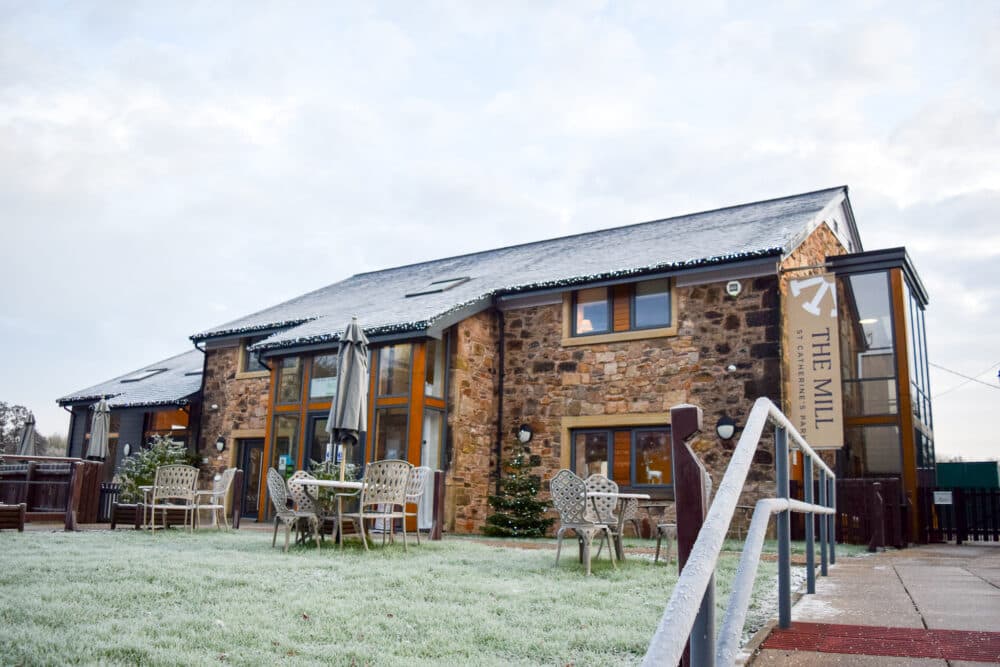 Stone building labeled "The Mill" with outdoor seating and light snow on the ground. - Home Instead