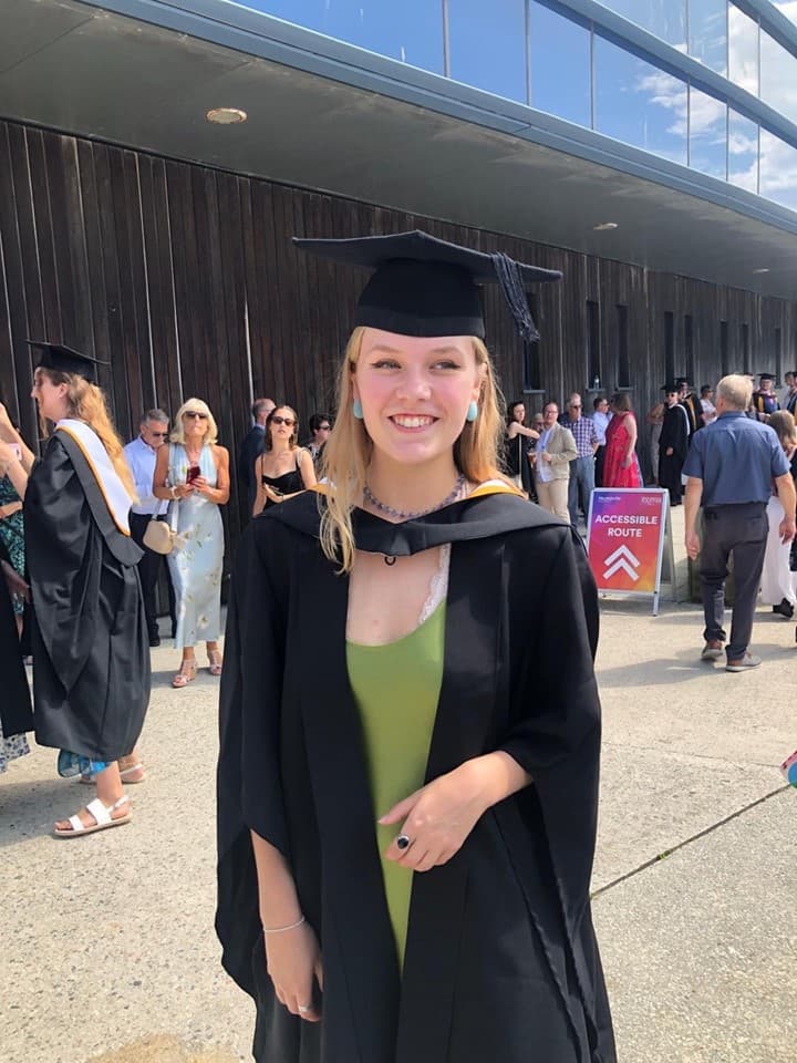 Graduate in cap and gown smiling at an outdoor ceremony with people in the background. - Home Instead