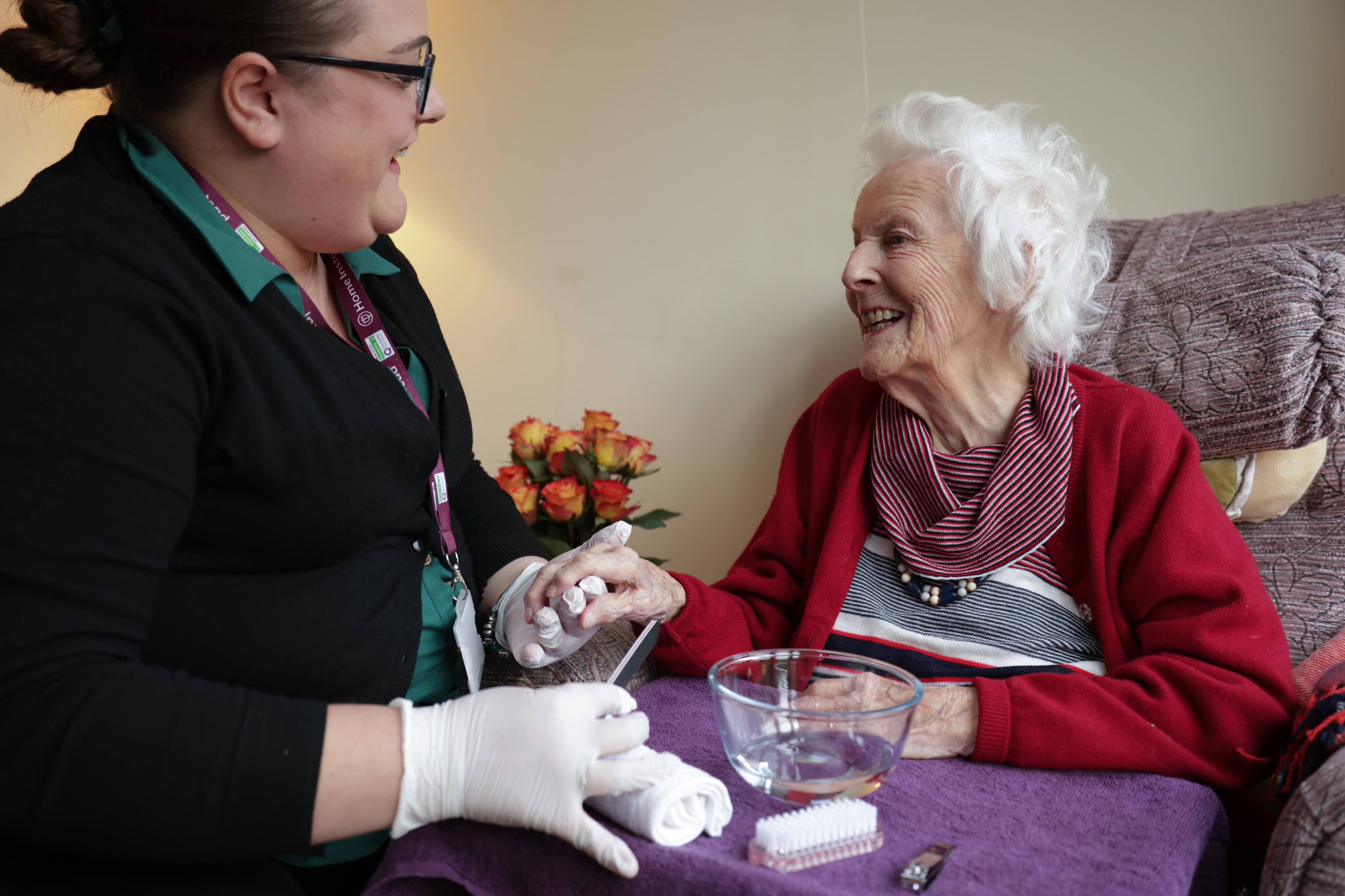A caregiver smiles while holding an elderly woman's hand during a manicure at home. - Home Instead