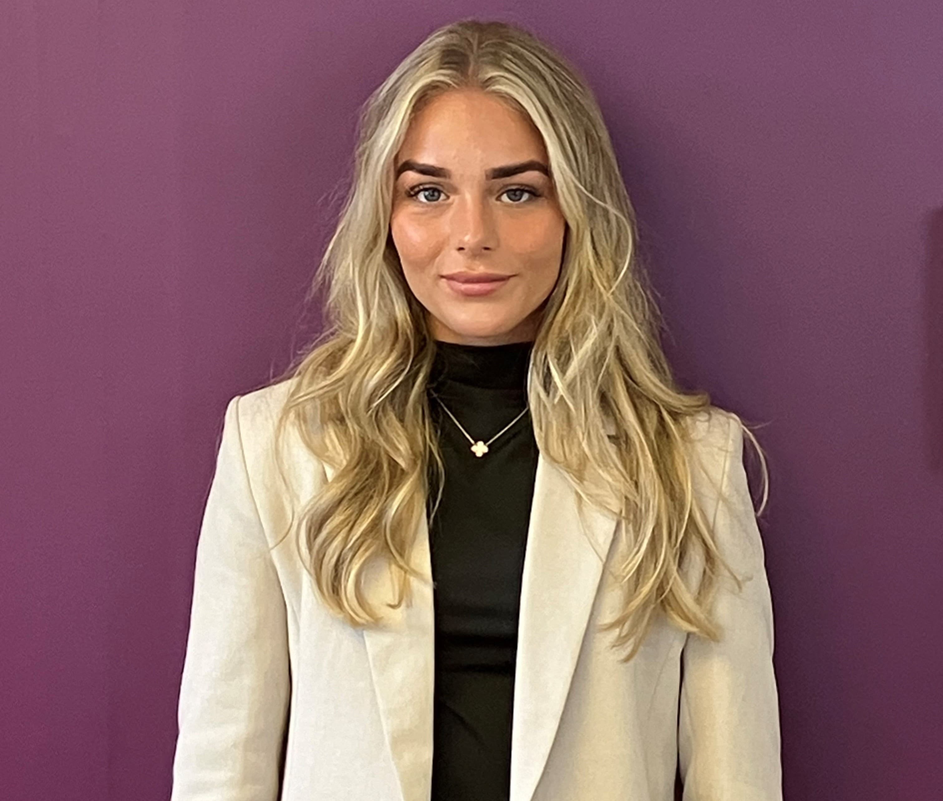 Woman with long blonde hair wearing a white blazer and black top, standing in front of a purple wall. - Home Instead