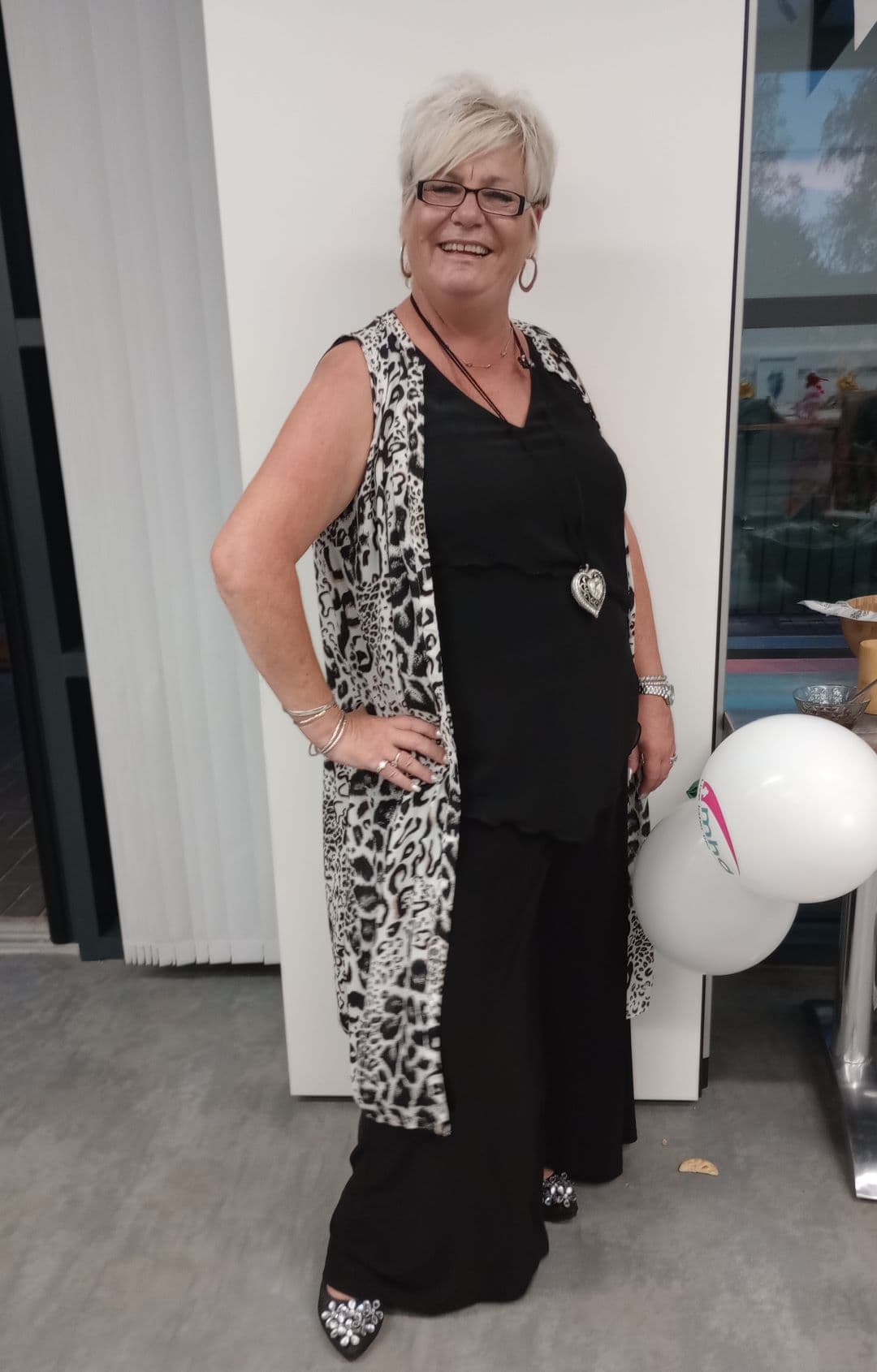 Smiling woman in black outfit and leopard print vest, posing indoors beside a balloon. - Home Instead
