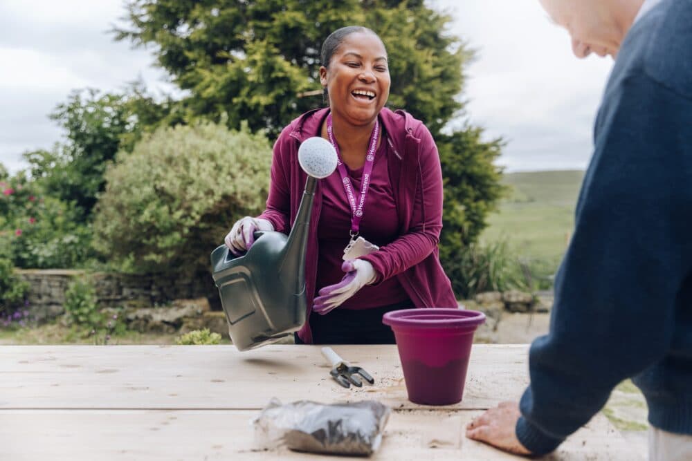Smiling care professional holding a watering can, watering a potted plant on an outdoor table alongside a client, fostering a moment of connection and well-being. - Home Instead York