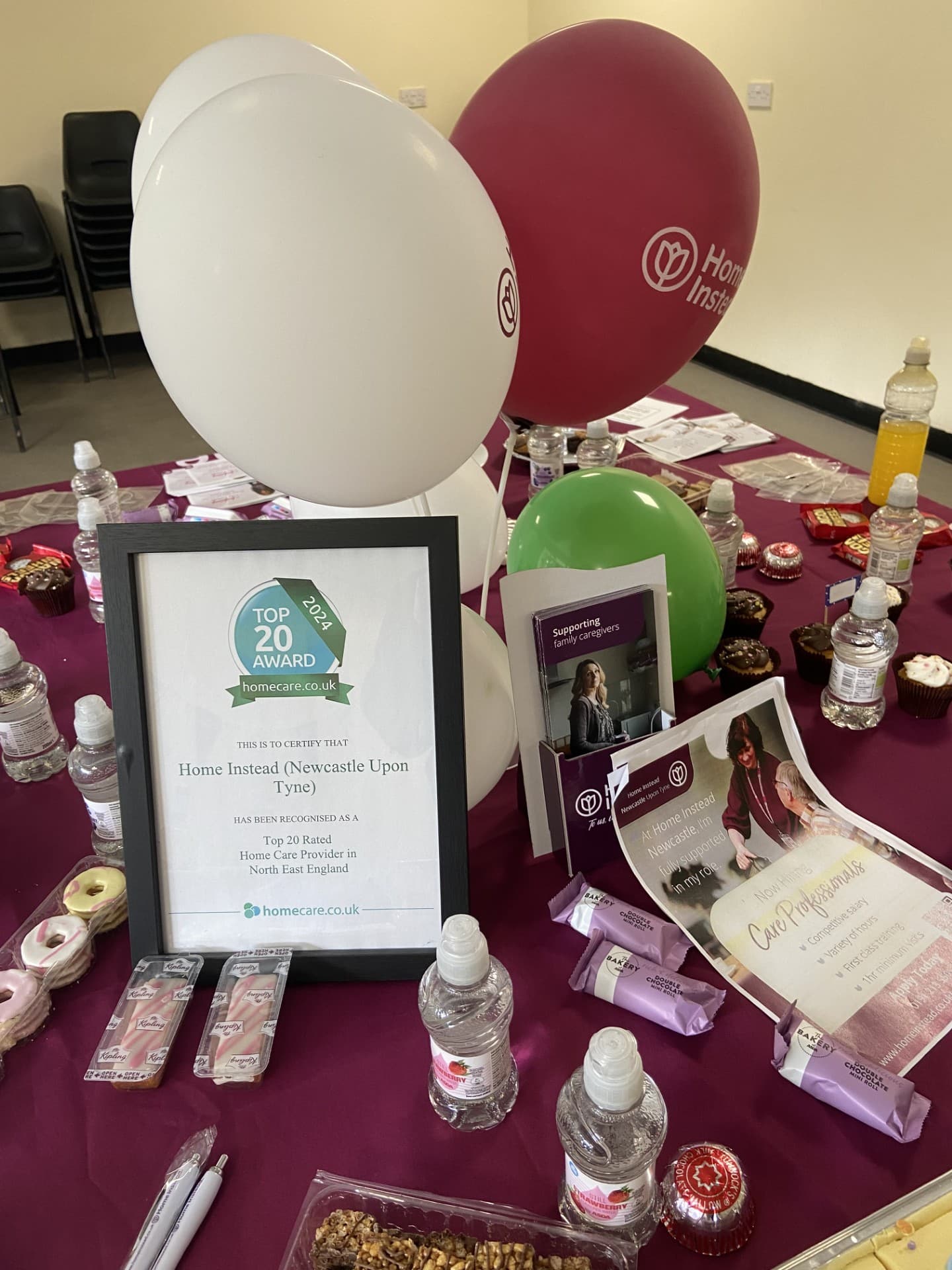 Award certificate on a table with branded balloons, snacks, water bottles, and promotional materials. - Home Instead