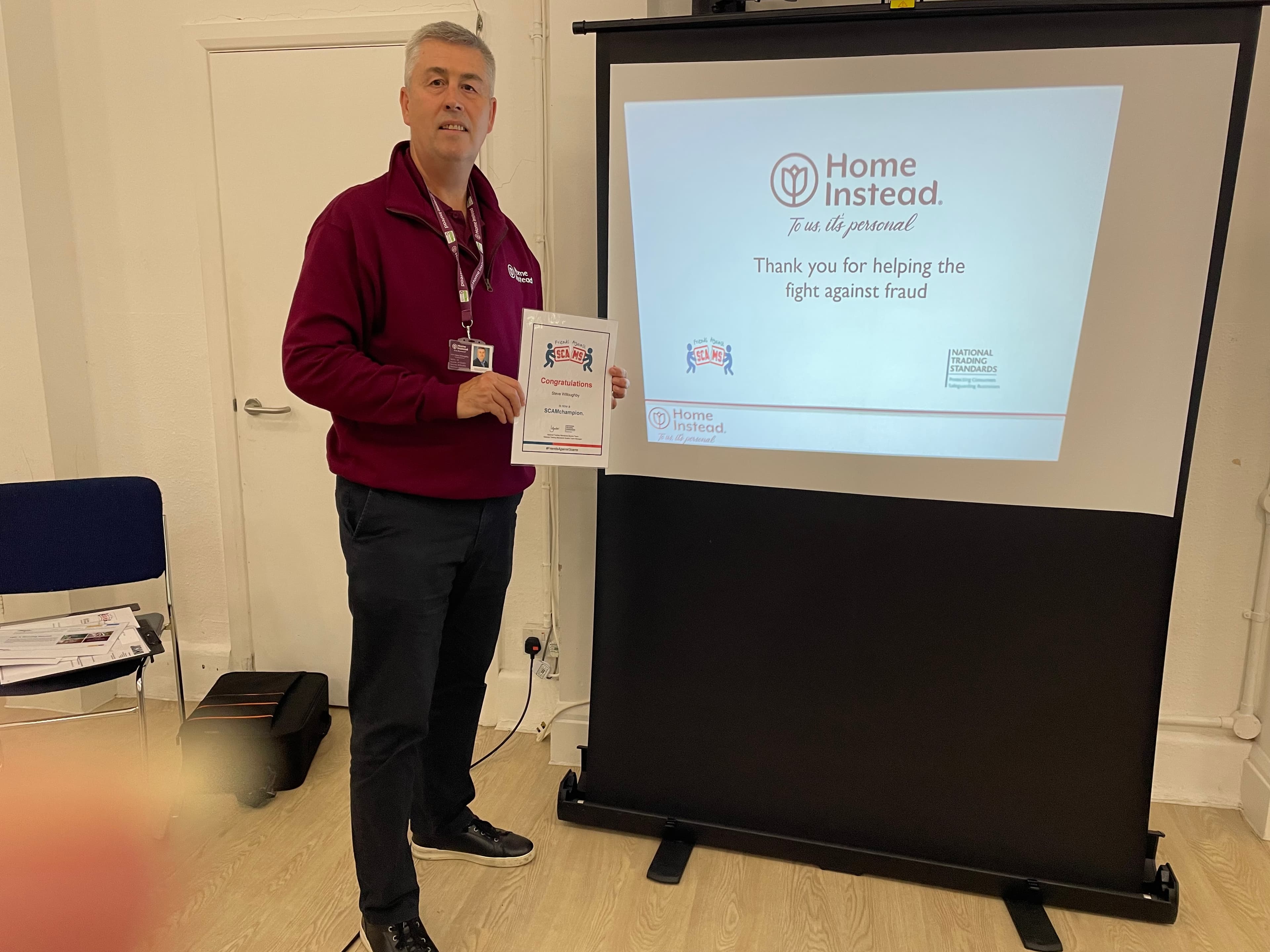Man holding a certificate stands beside a screen displaying a Home Instead anti-fraud message. - Home Instead