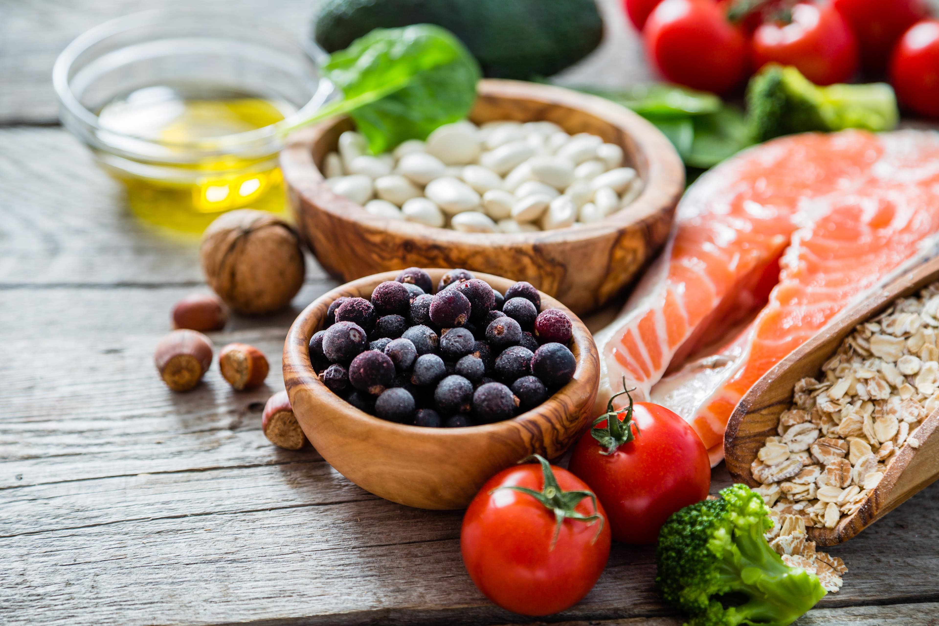 Variety of healthy foods including salmon, beans, berries, tomatoes, nuts, oats, broccoli, and olive oil on a wooden table. - Home Instead