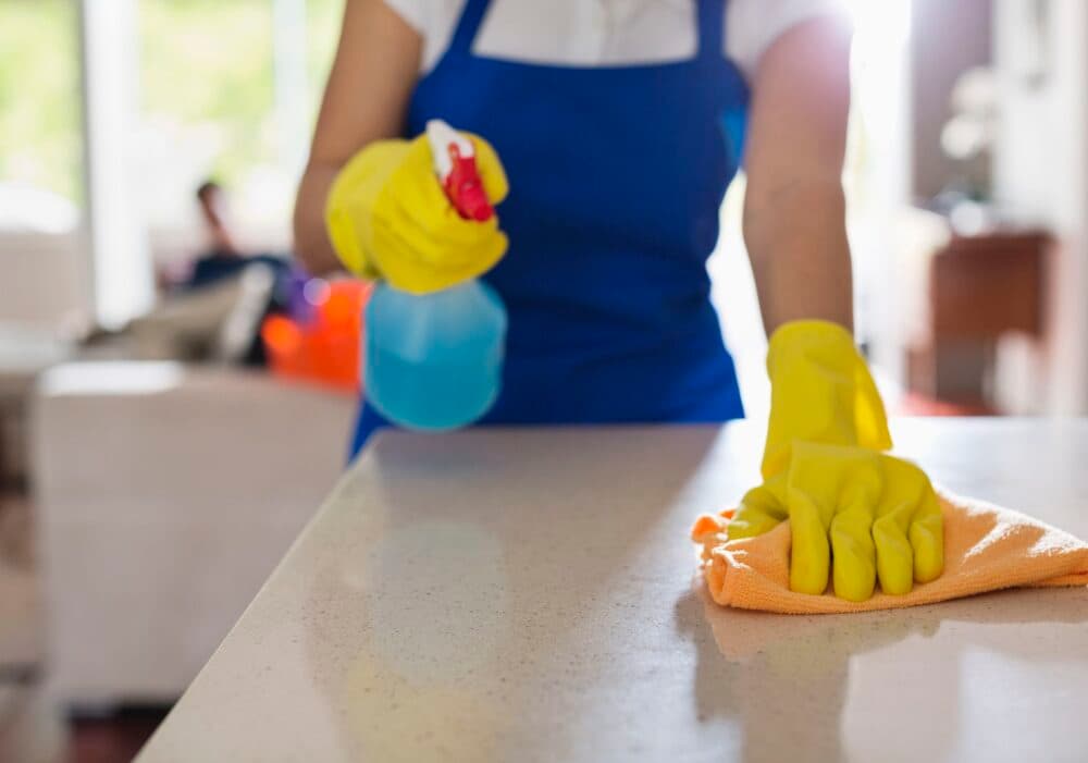 Person wearing yellow gloves cleaning a kitchen counter with a spray bottle and orange cloth. - Home Instead