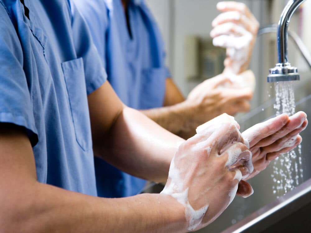 Two people in scrubs washing their hands with soap at a sink, with water running. - Home Instead