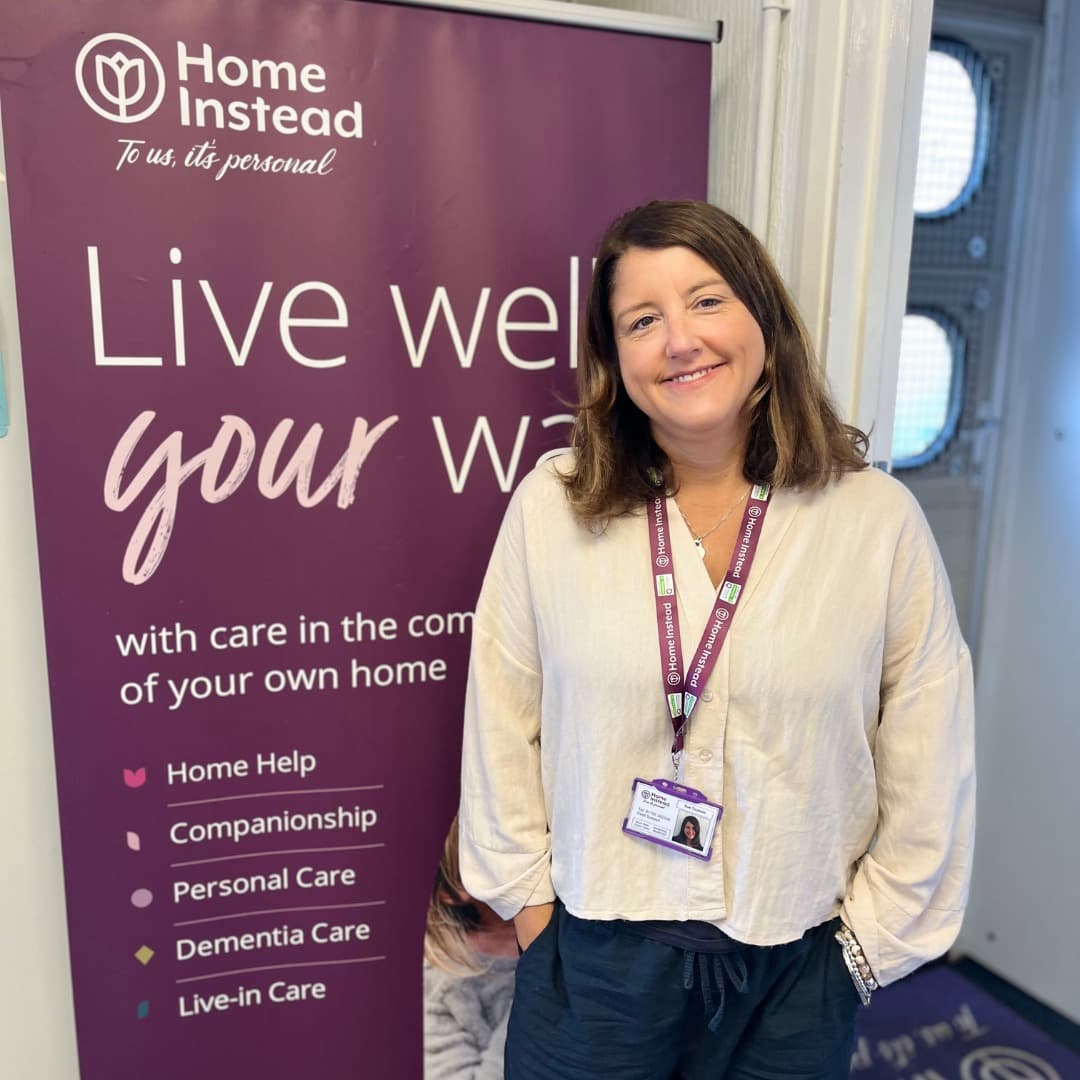 Image of Sue a new member of our Field Support Team. Sue has big smile on her face and is standing in the office in front of the Live Well your Way banner.