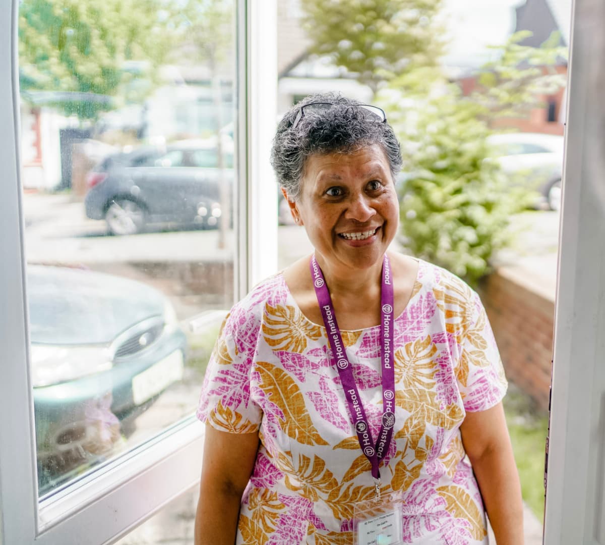 A lady at the door wearing printed shirt with short grey hair and wearing an ID lanyard smiling while entering the door