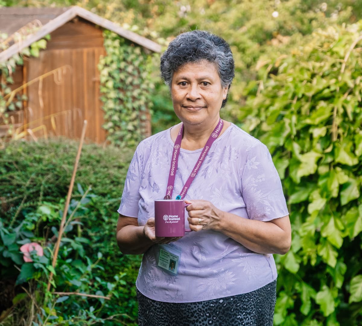 A person holds a mug, standing in a garden with a wooden shed and green plants in the background. - Home Instead