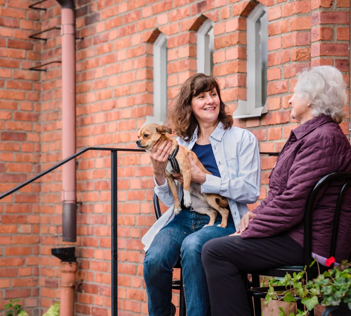 A senior woman with white hair wearing winter clothe outside of the house while speaking to a youner woman wearing long sleeves and holding a dog