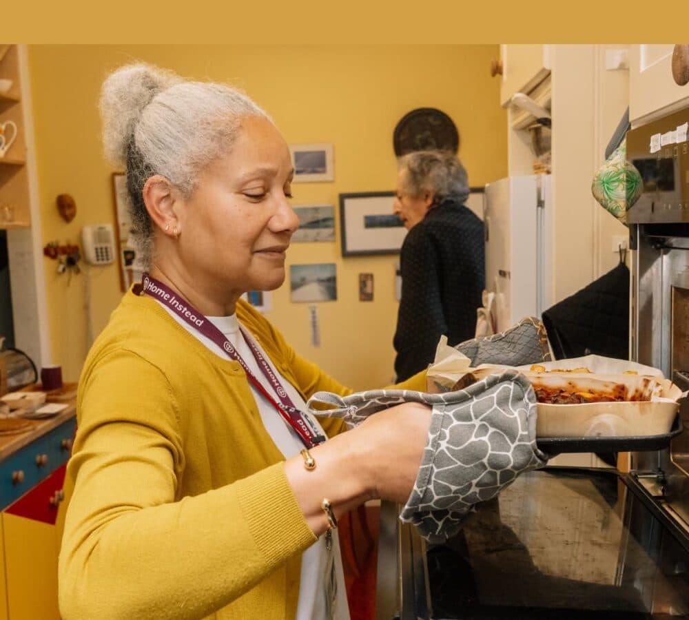 A lady wearing mistard sweater with grey hair holding a pan and baking a dish inside the kitchen