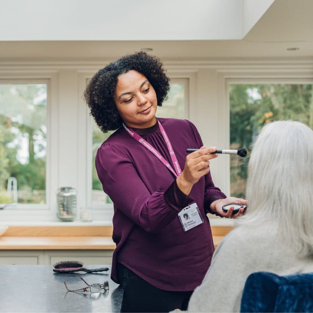 A woman in a purple shirt applies makeup to an elderly person in a bright room. - Home Instead