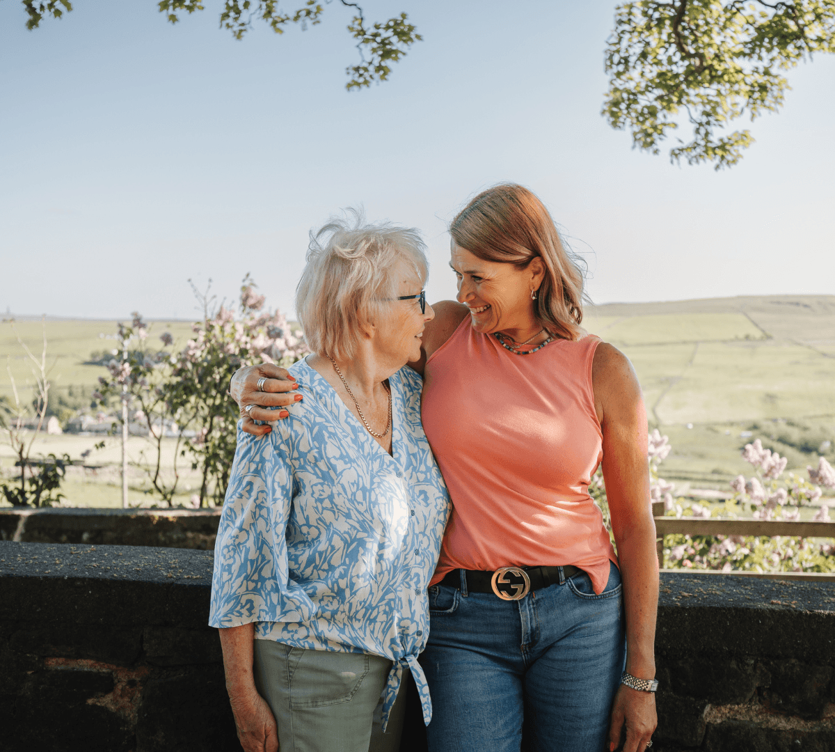 A Senior woman wearing eyeglasses and blue blouse with short hair looking at a younger woman who is wearing peach top and pants standing outside with a huge field at the background