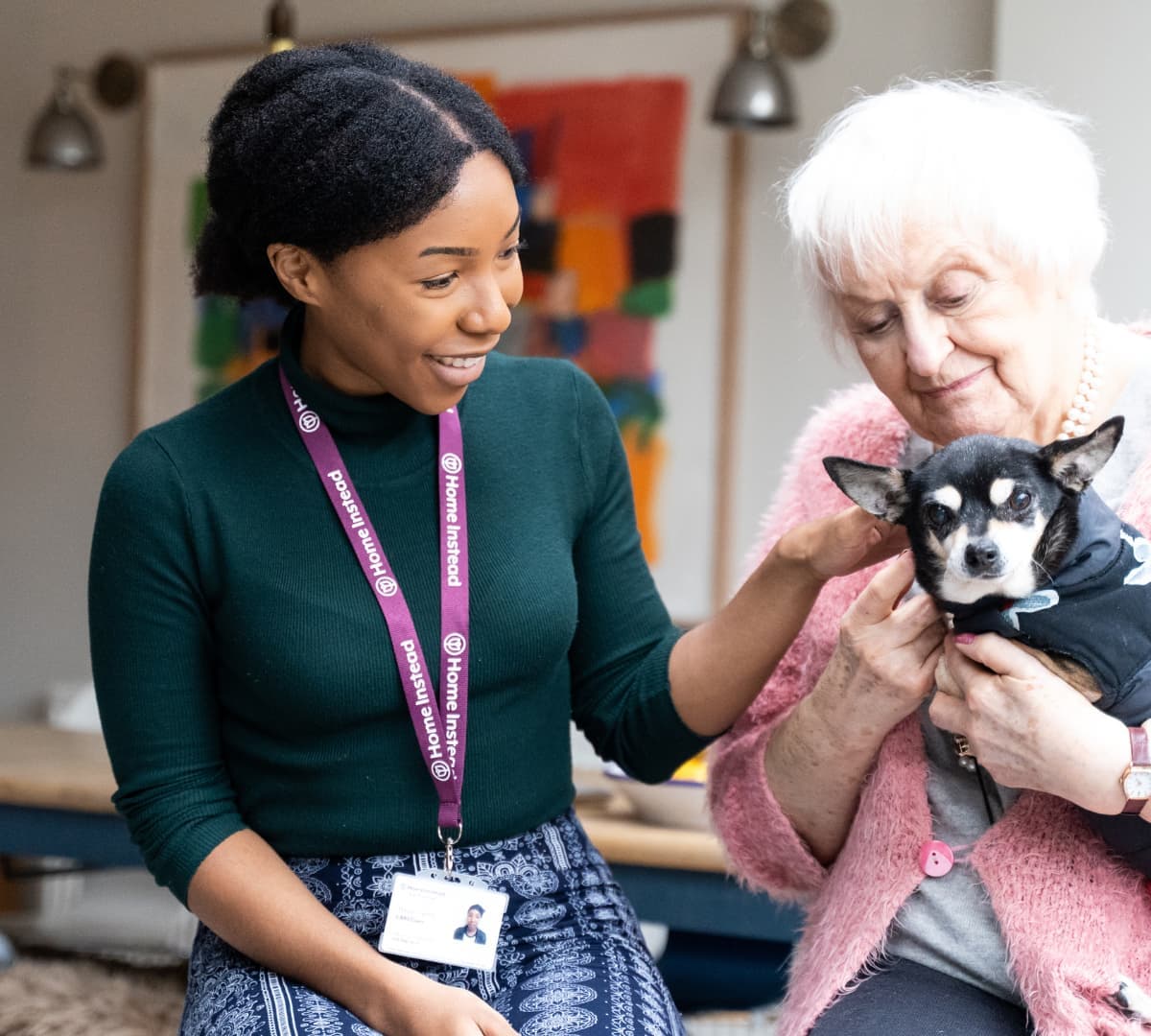 Senior woman with white hair smiling while holding a dog with her younger carer with black hair and smiling inside the house