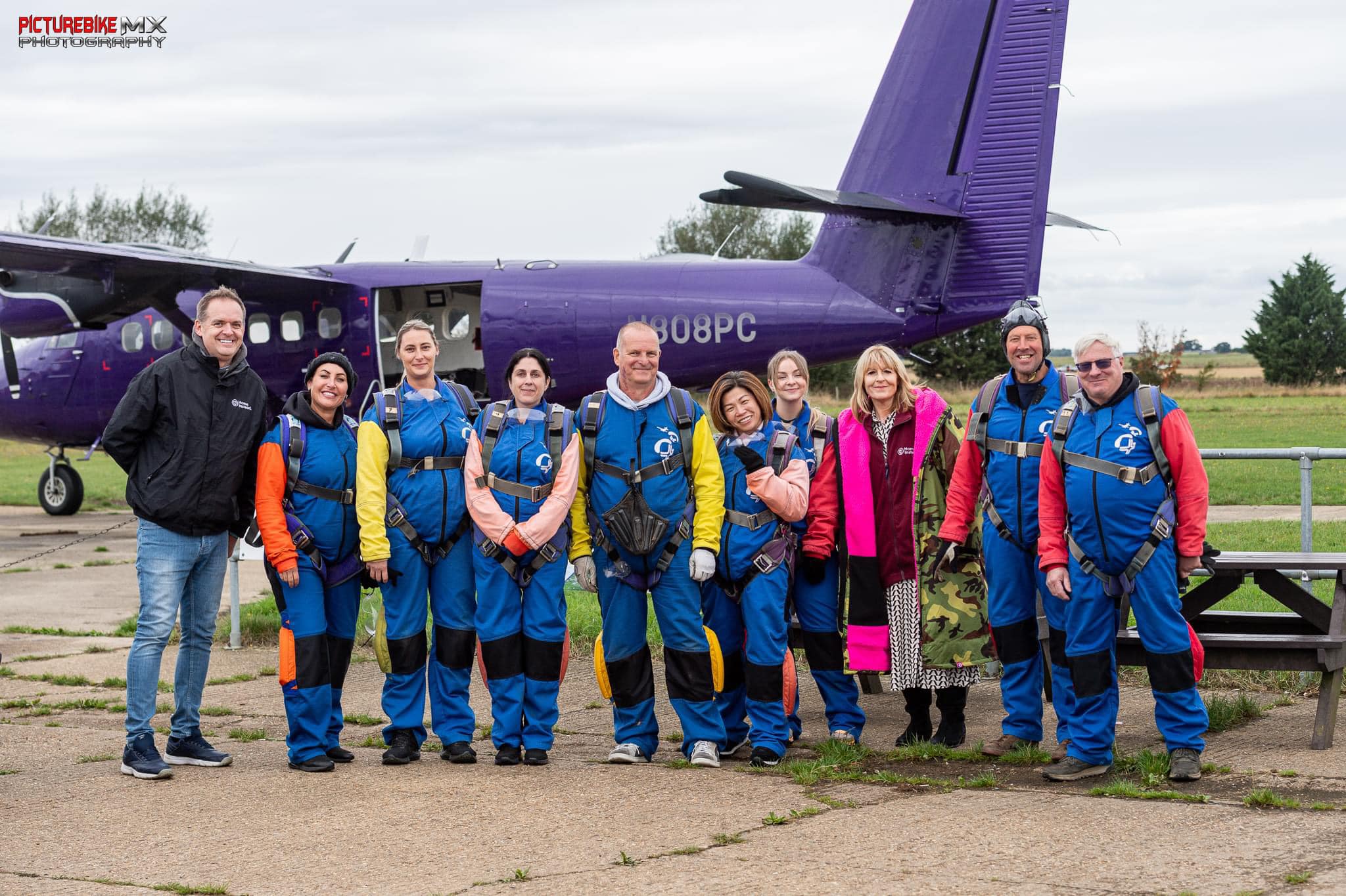 A group of people in jumpsuits stands in front of a purple airplane, smiling at the camera. - Home Instead