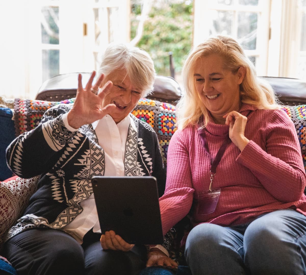 A senior woman with her carer sitting on a couch and near the window while holding a device and smiling and waving