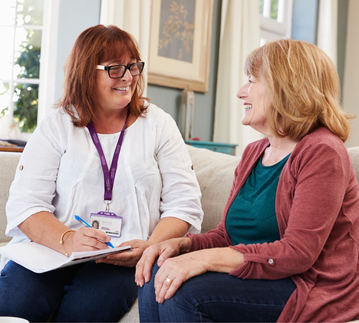 Two woman looking at each other smiling and talking while sitting on a couch