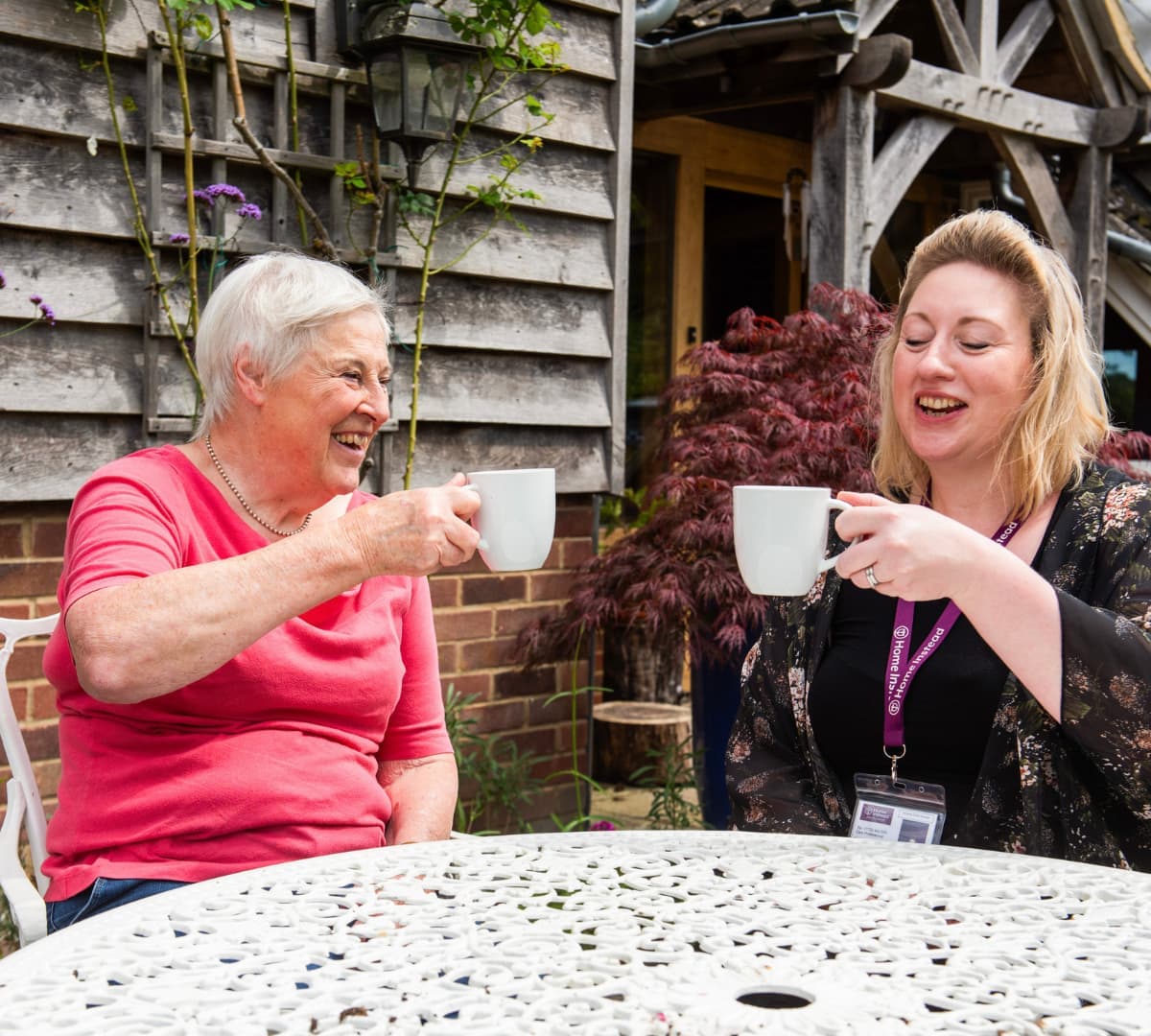 A woman with black top holding her cup while smiling and sitting on a chair with a senior woman with grey hair smilign and holding her cup up while sitting on a chair outside the house