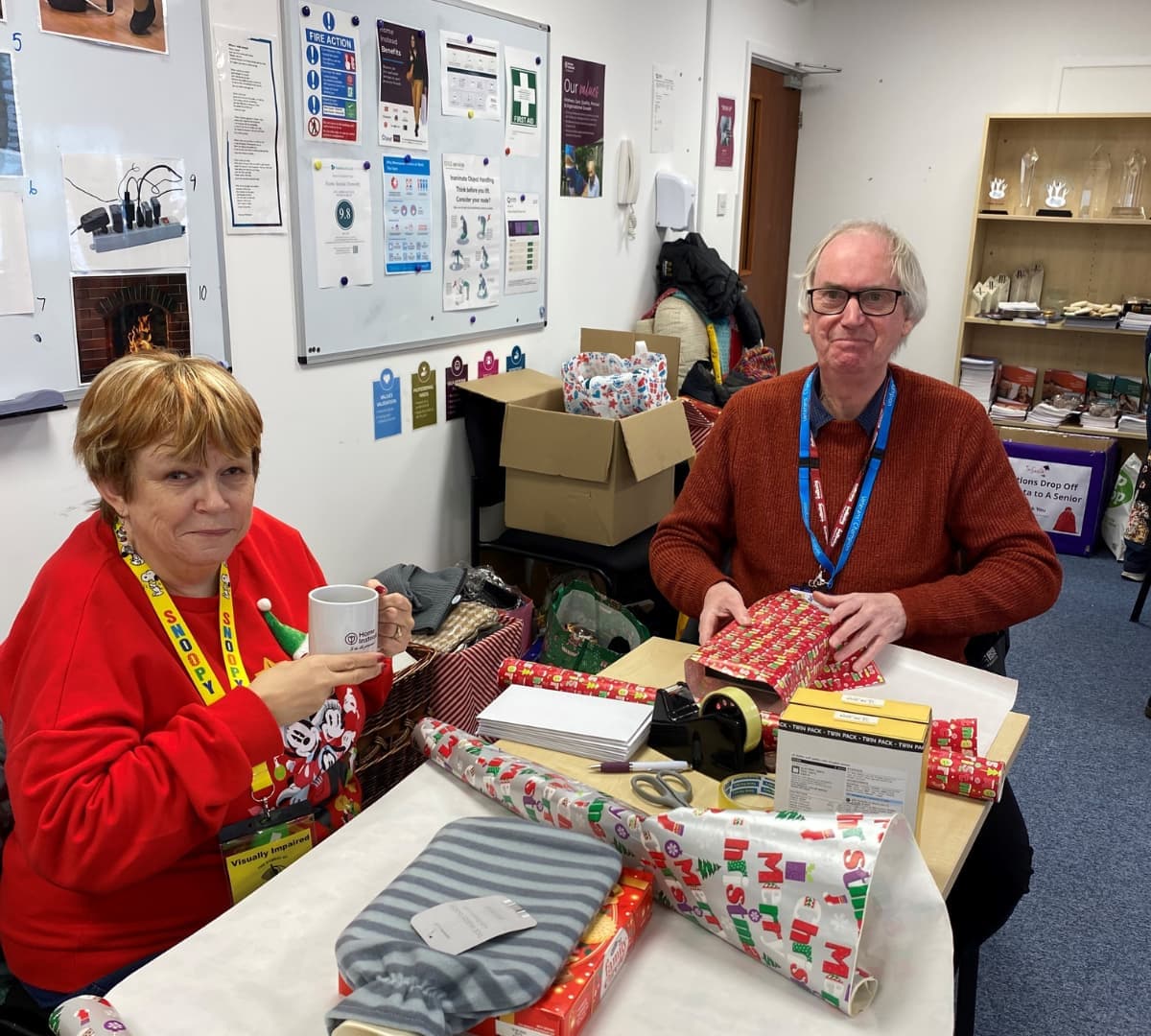 An older man smiling while wrapping gifts and older woman holding a cup inside an office room