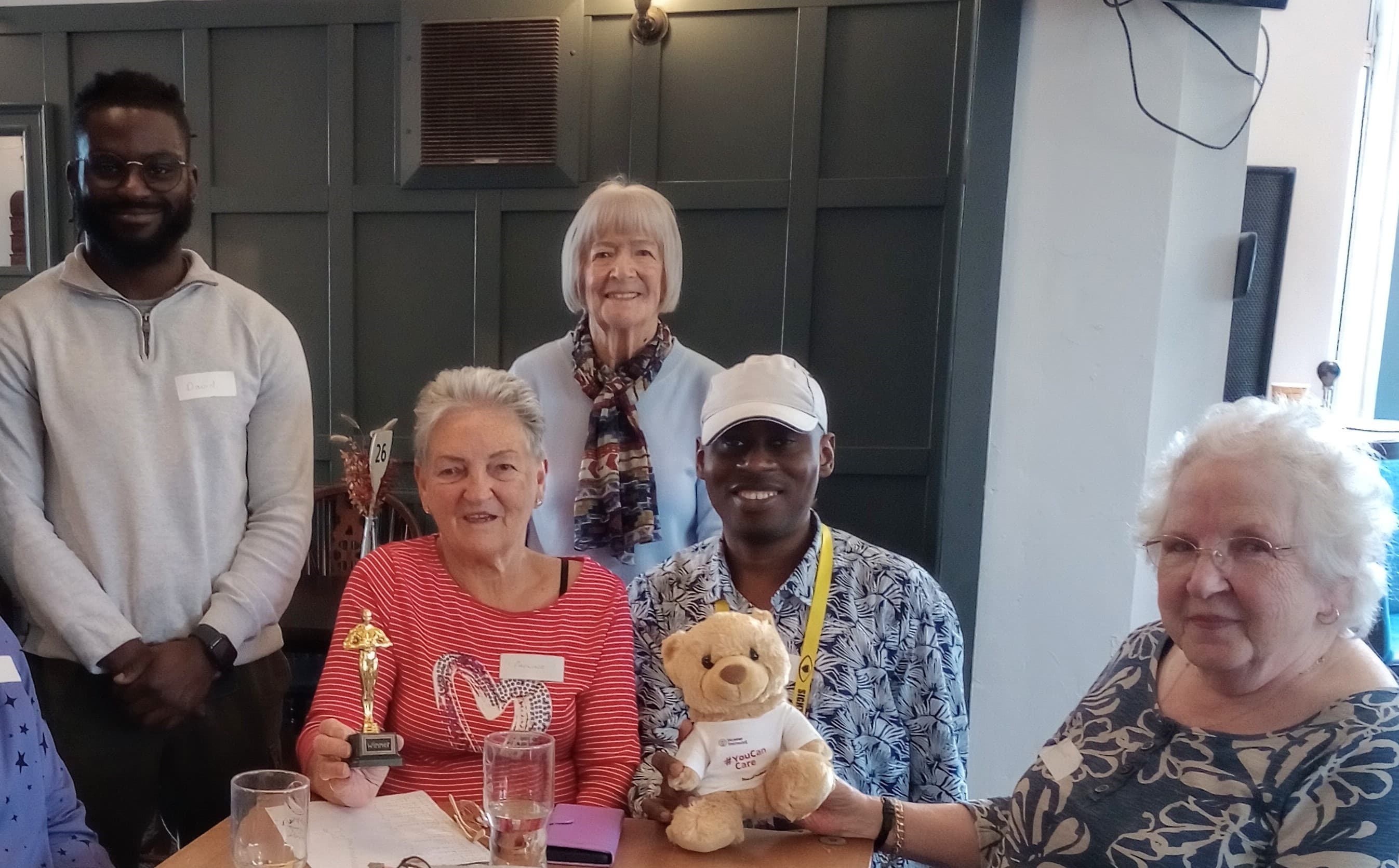 Five people smiling at a table, one holding a trophy and another holding a teddy bear with a t-shirt. - Home Instead