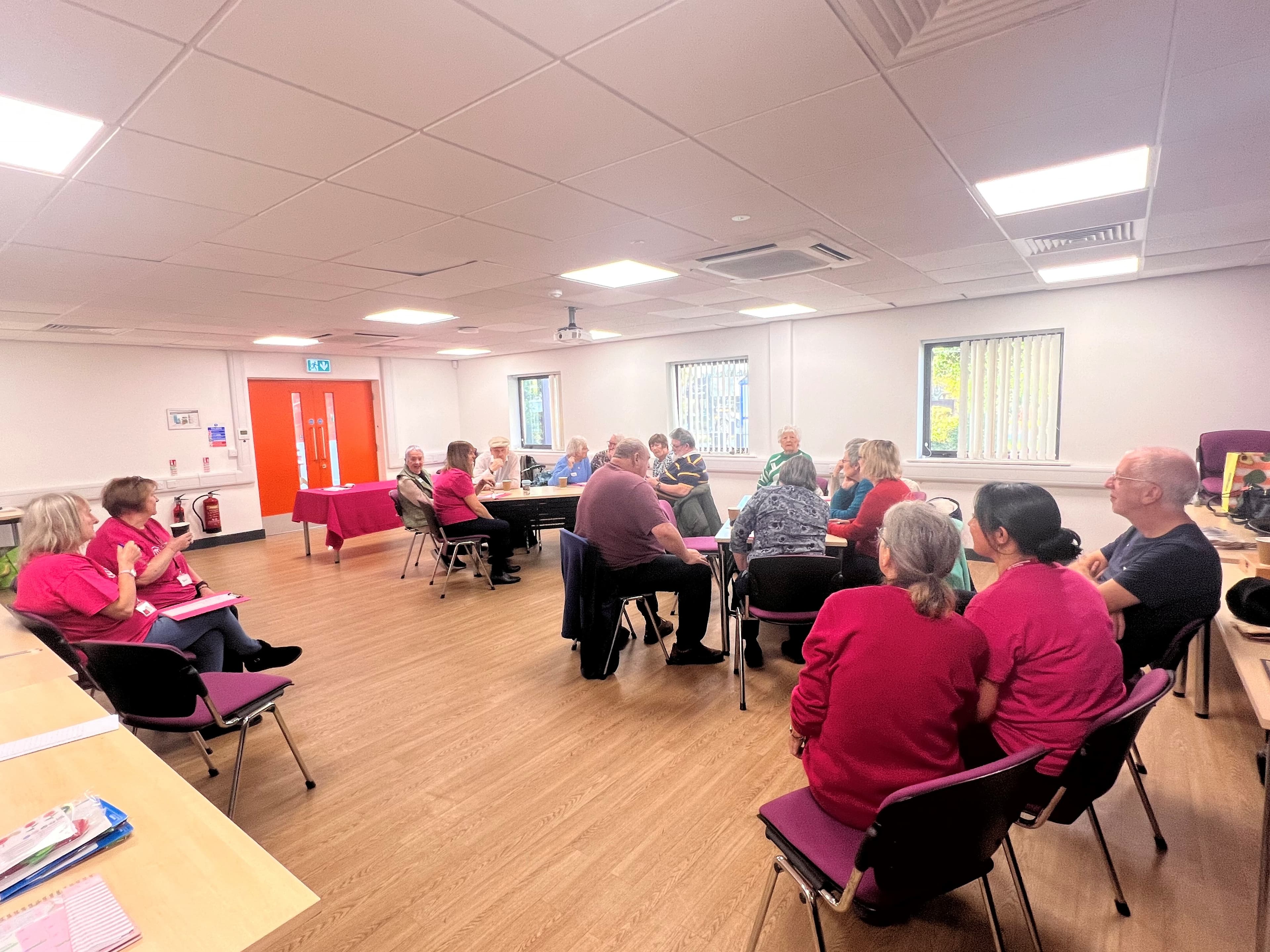 A group of adults sit in a bright room, gathered around tables for a meeting or discussion. - Home Instead
