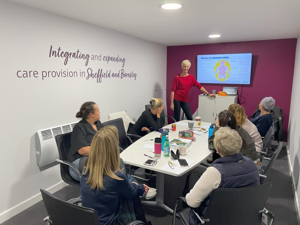 A woman presents to six people in a meeting room with a health-related slide displayed on a screen. - Home Instead
