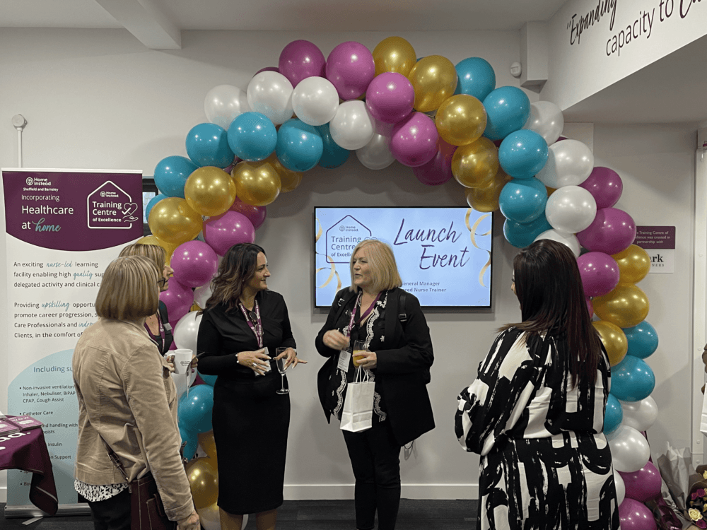 Four women talk beneath a balloon arch at a launch event, with a screen displaying "Launch Event" behind them. - Home Instead