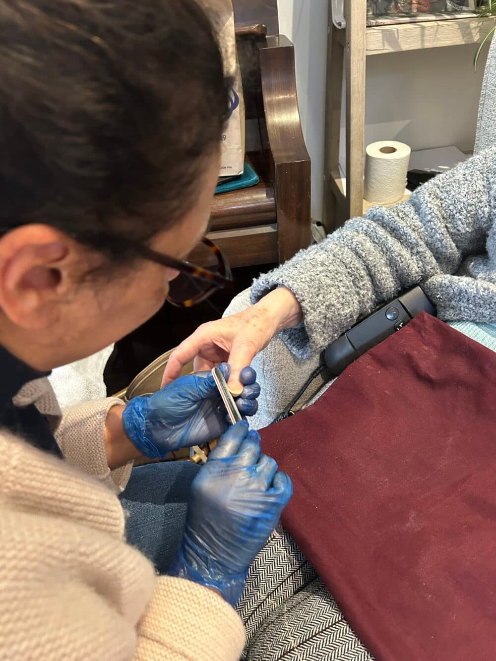 A person in blue gloves files another person's nails while they rest their arm on a burgundy cloth. - Home Instead