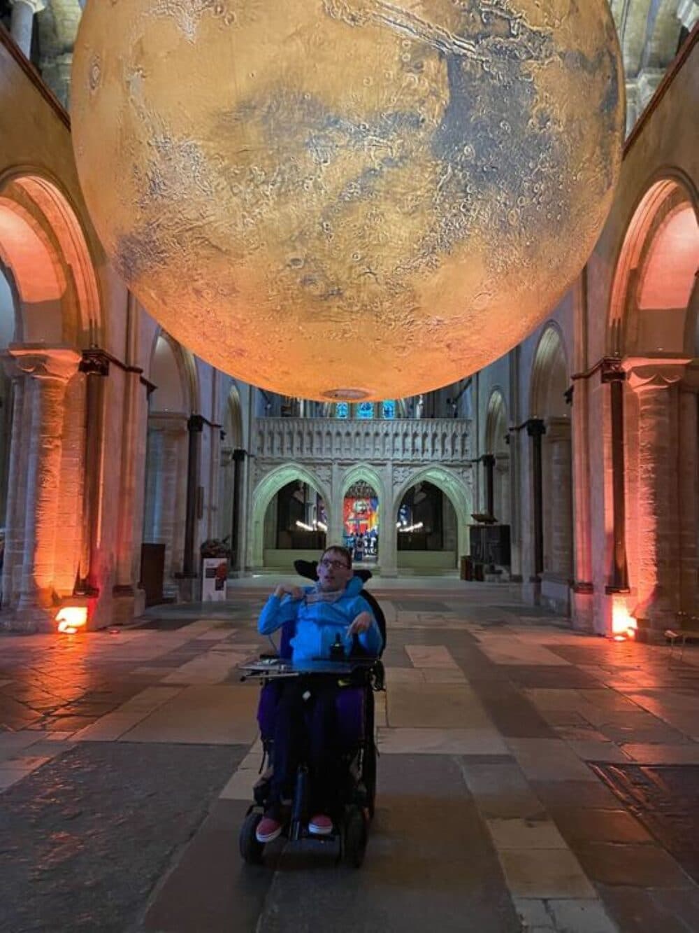 Person in a wheelchair beneath a large model of the moon in a grand, arched cathedral interior. - Home Instead