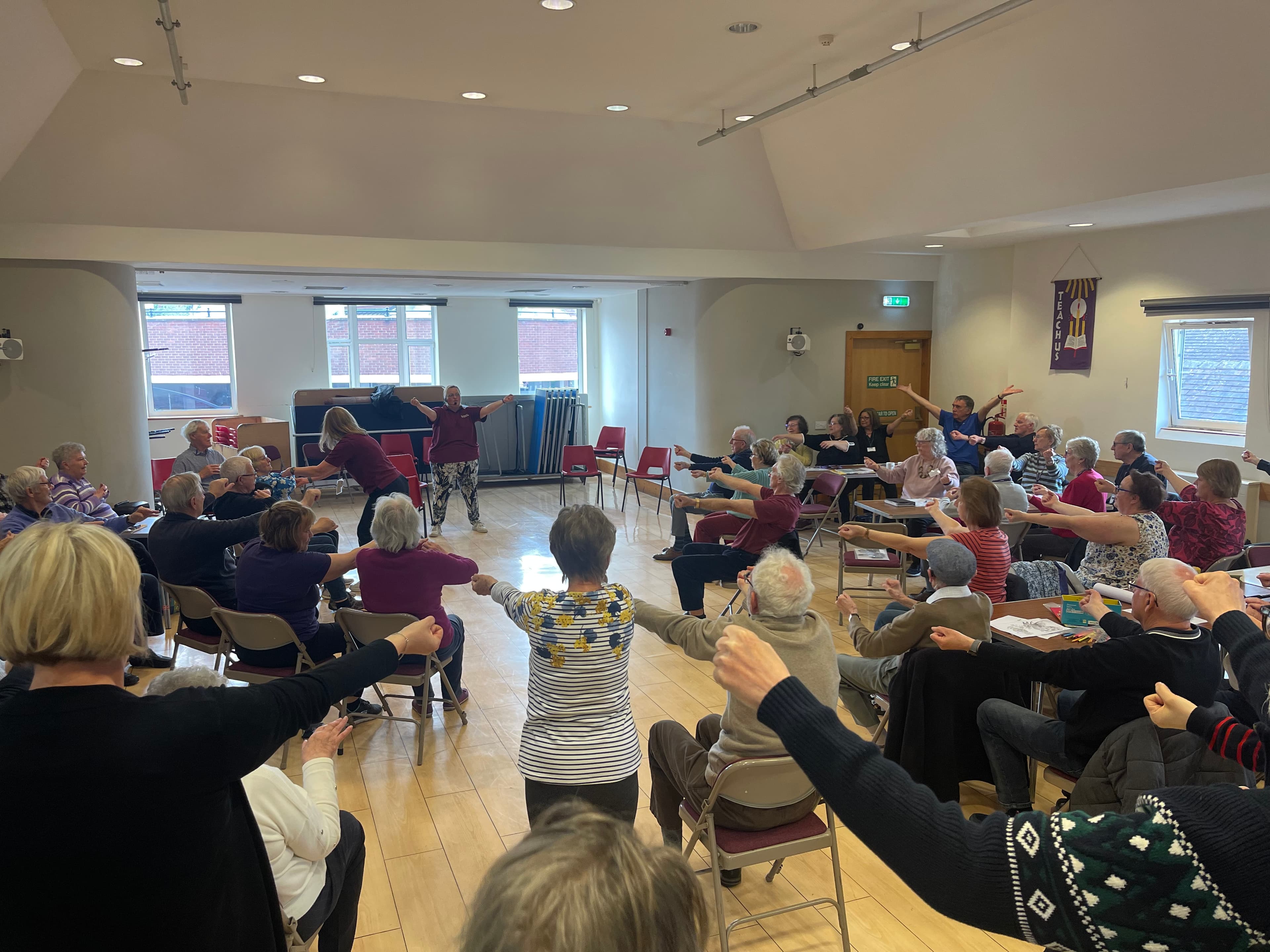 A group of older adults sit in a circle, raising their arms together in a well-lit community room. - Home Instead