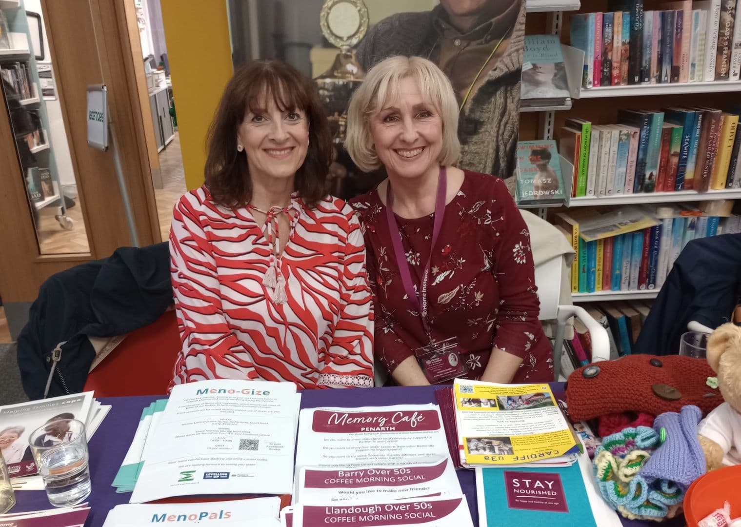 Two smiling women sit at a table with leaflets about social events and health in a library setting. - Home Instead