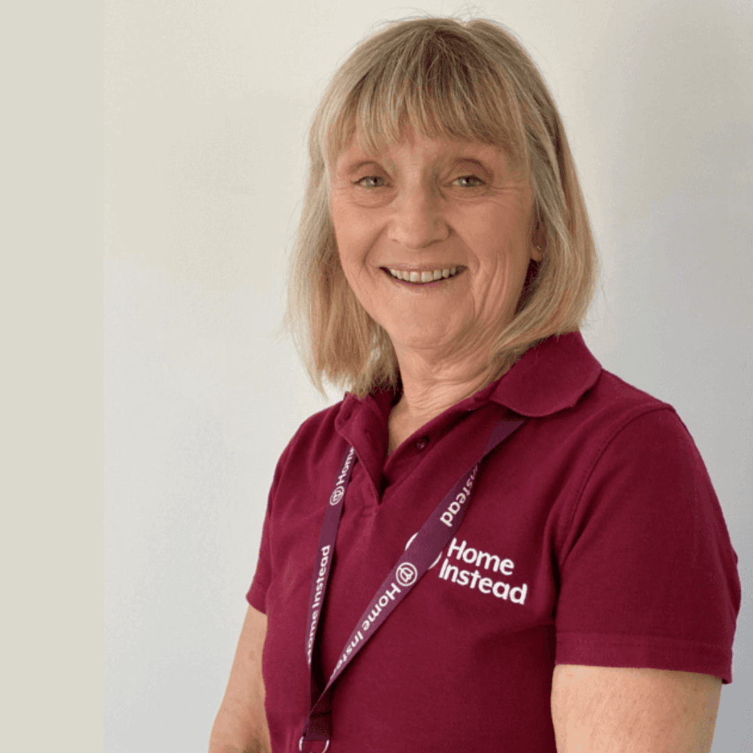 Smiling woman in a maroon "Home Instead" polo shirt and lanyard, standing against a light background. - Home Instead