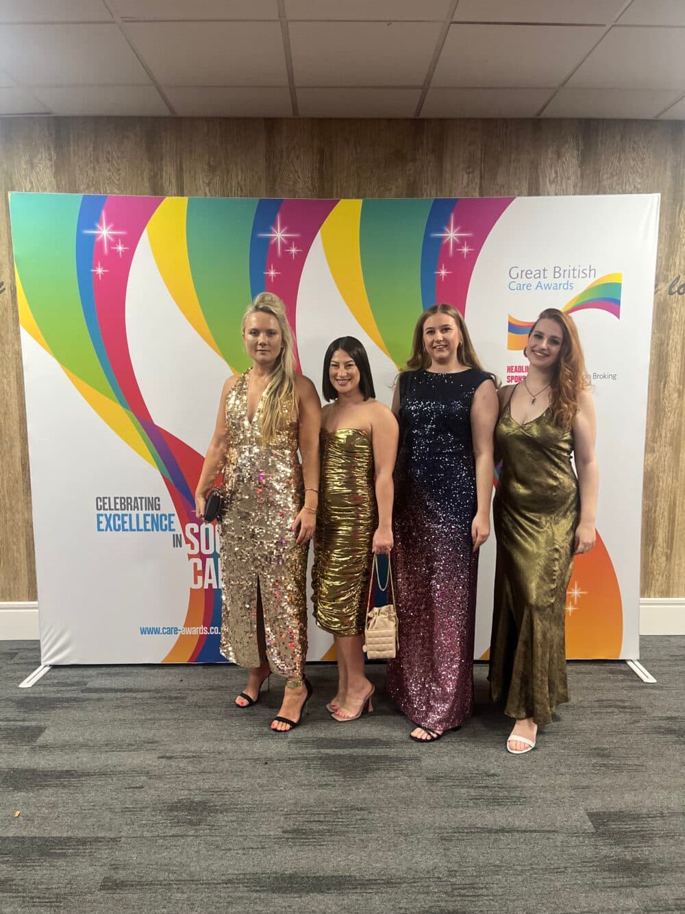 Four women in sparkly dresses stand in front of a colorful Great British Care Awards backdrop. - Home Instead