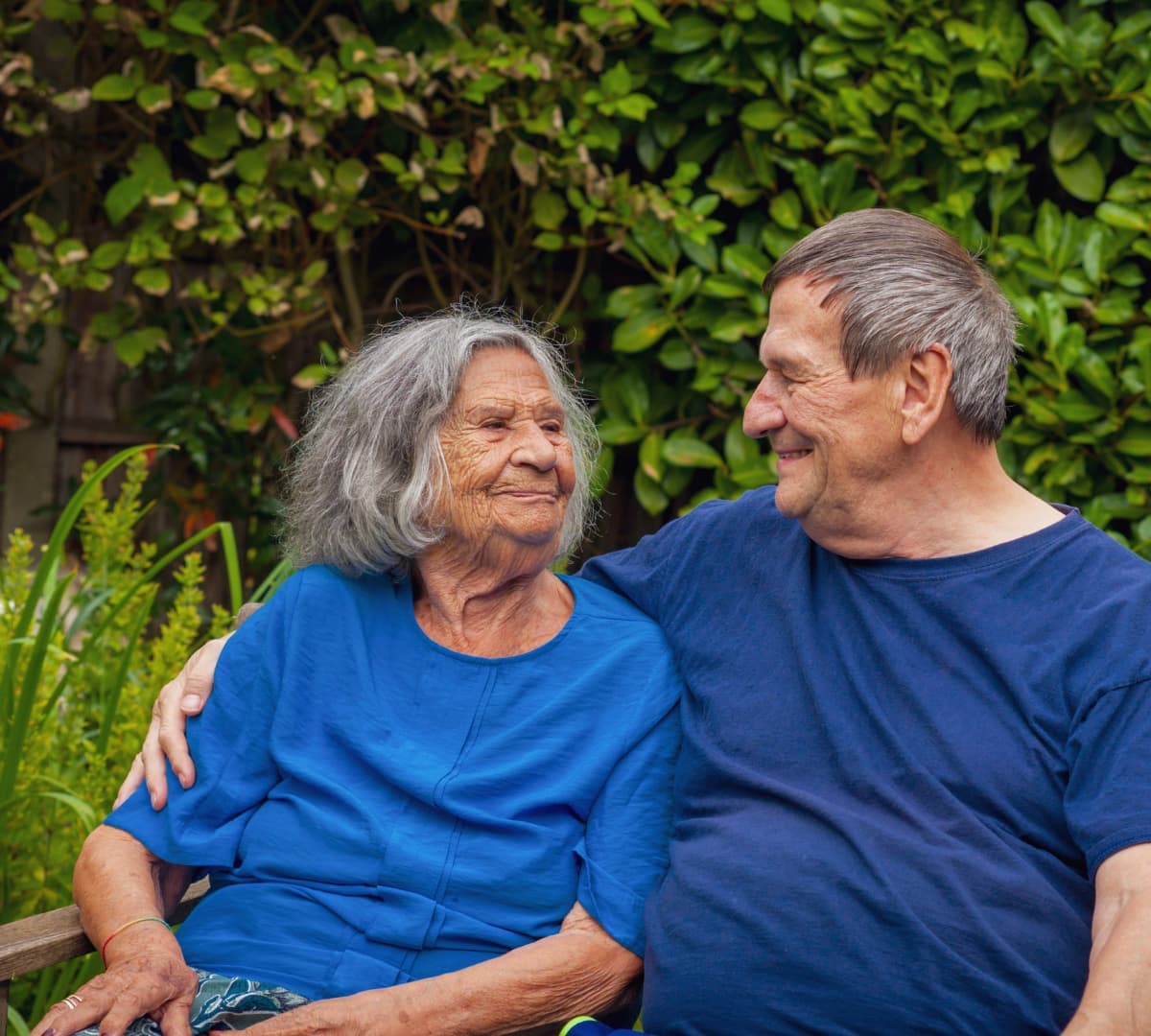 A senior mum and her son both with grey heair both wearing blue and sitting on a bench, happy and smiling outside the garden