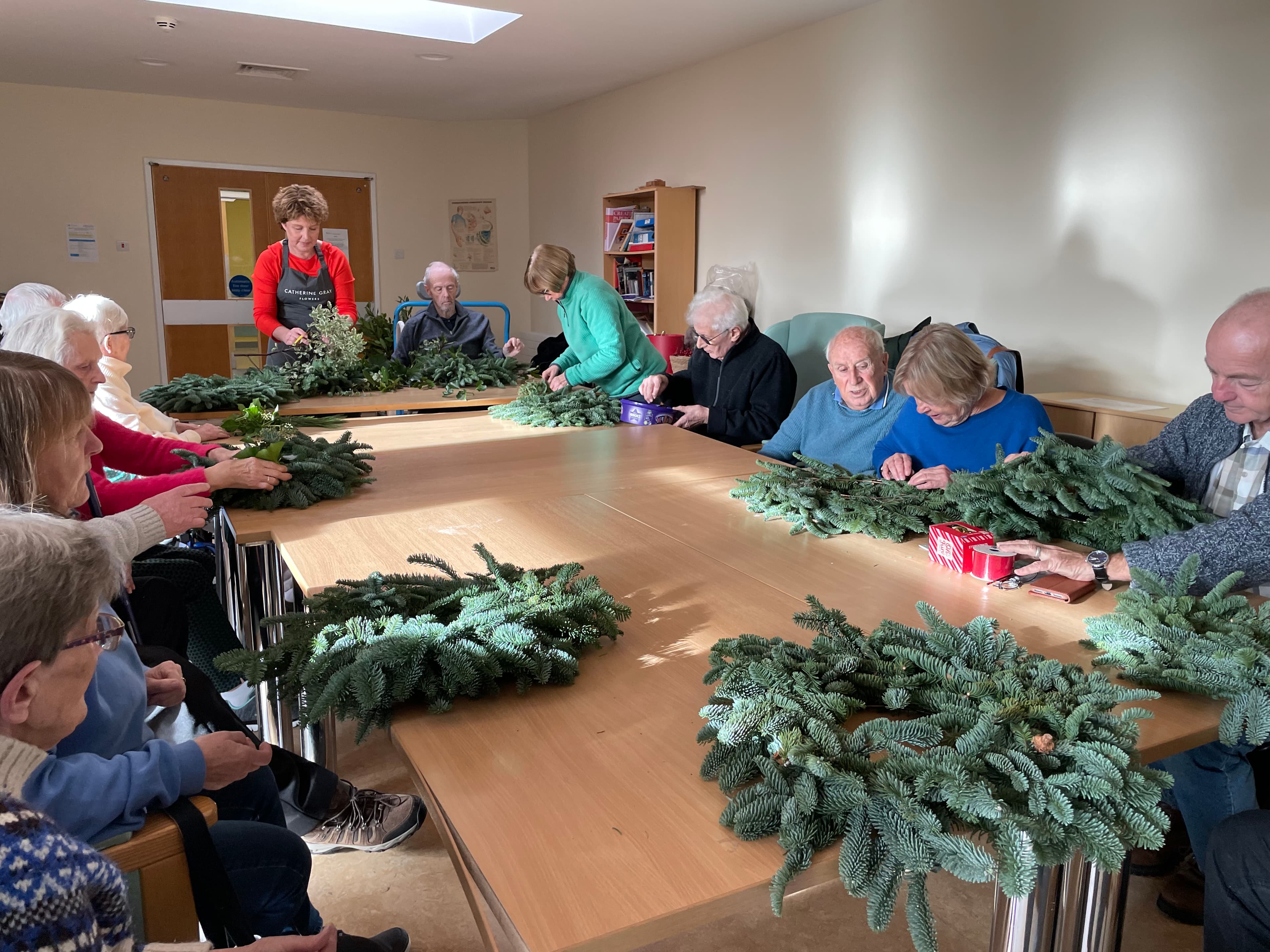 A group of seniors make festive wreaths together at a long table in a bright room. - Home Instead