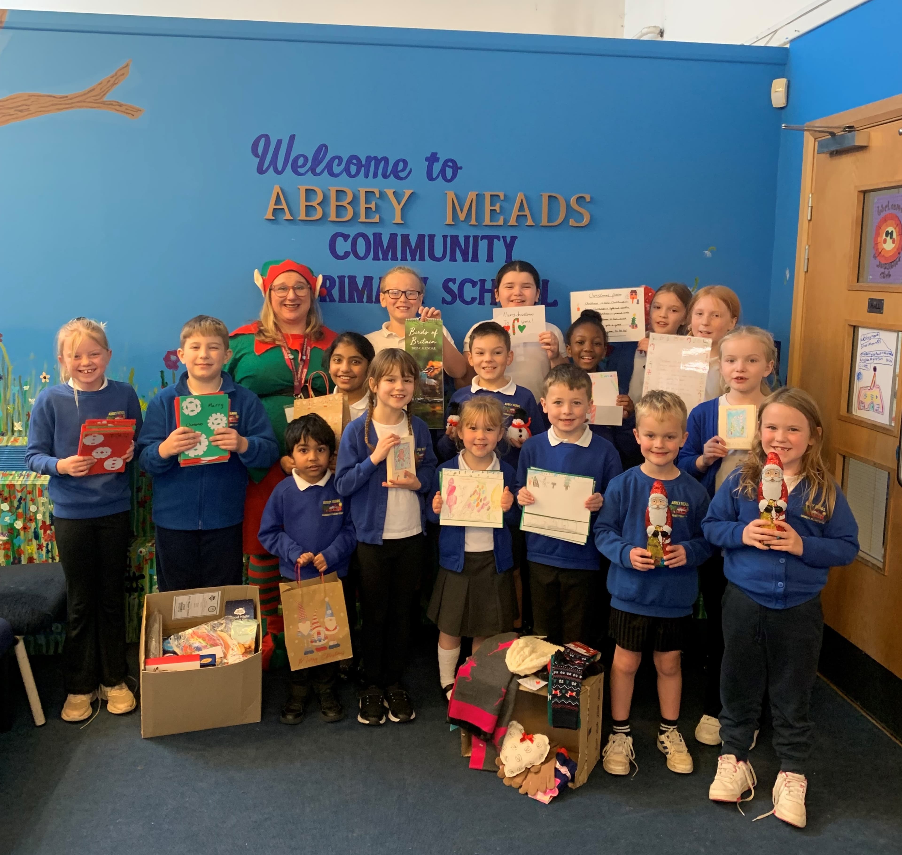 A group of children and an adult stand smiling with cards and gifts in front of a school welcome sign. - Home Instead
