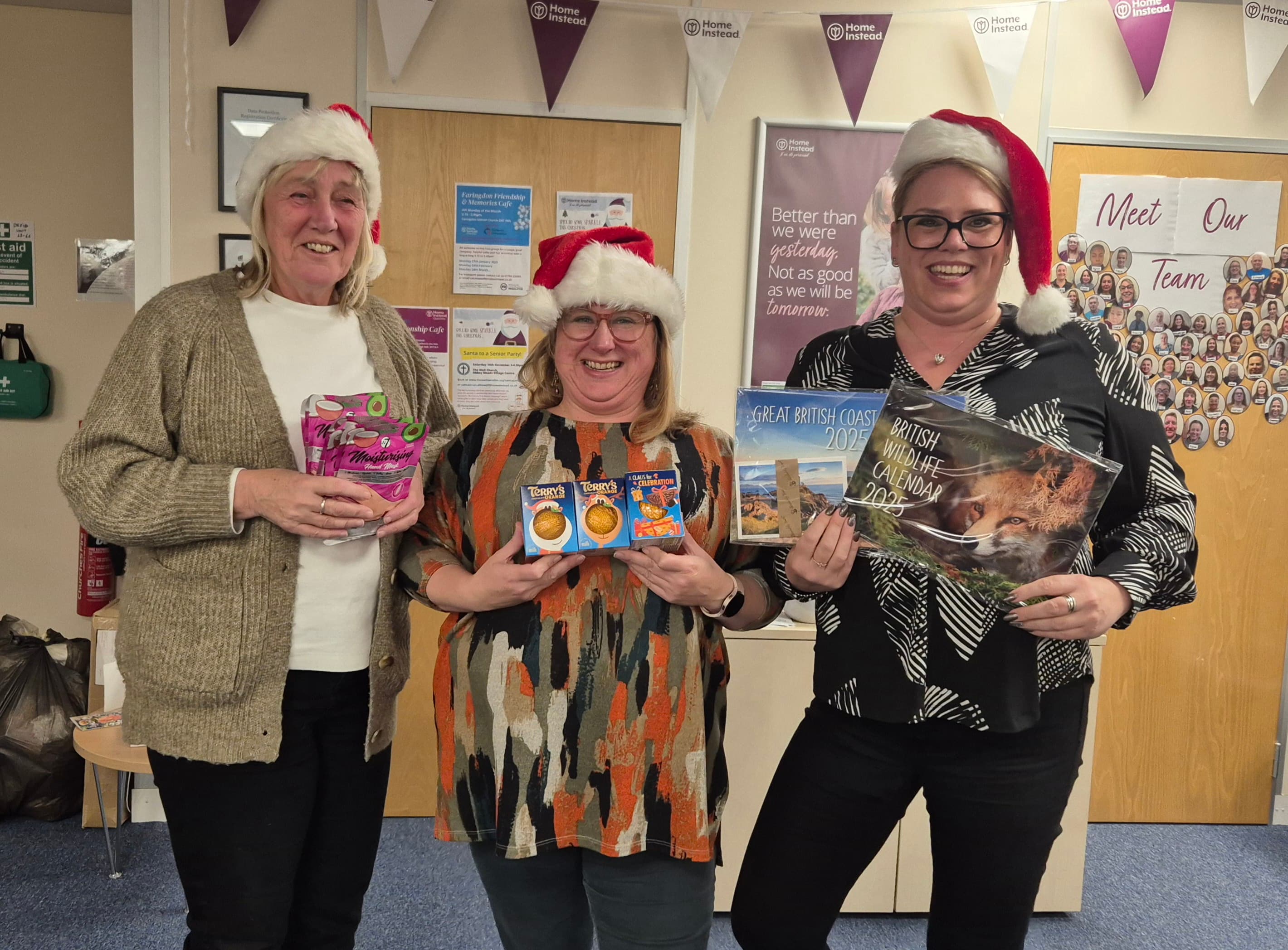 Three smiling women in Santa hats hold gifts while standing together in a festive office setting. - Home Instead