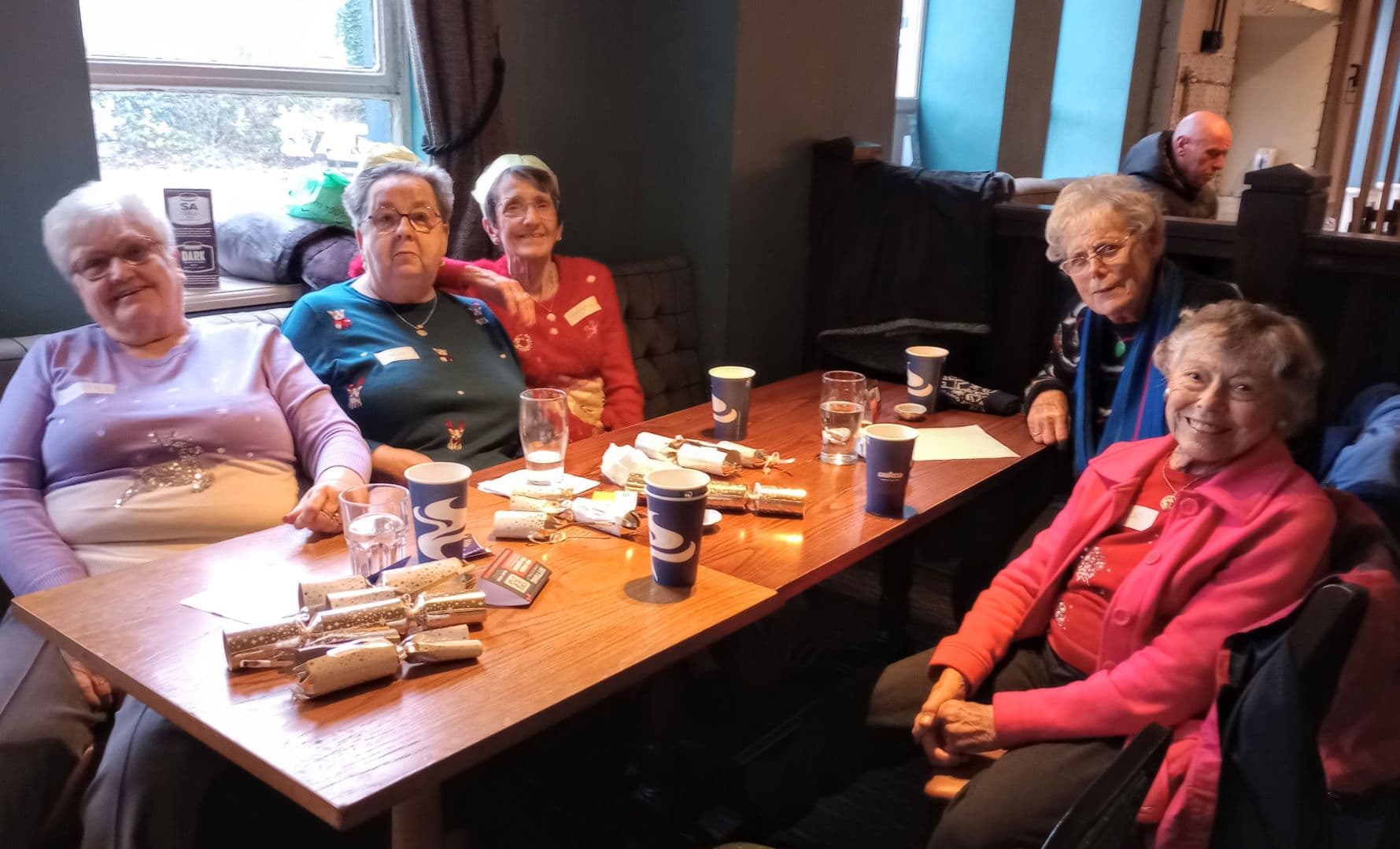 Five elderly women sit around a wooden table in a cozy cafe, smiling with cups, water glasses, and crackers. - Home Instead