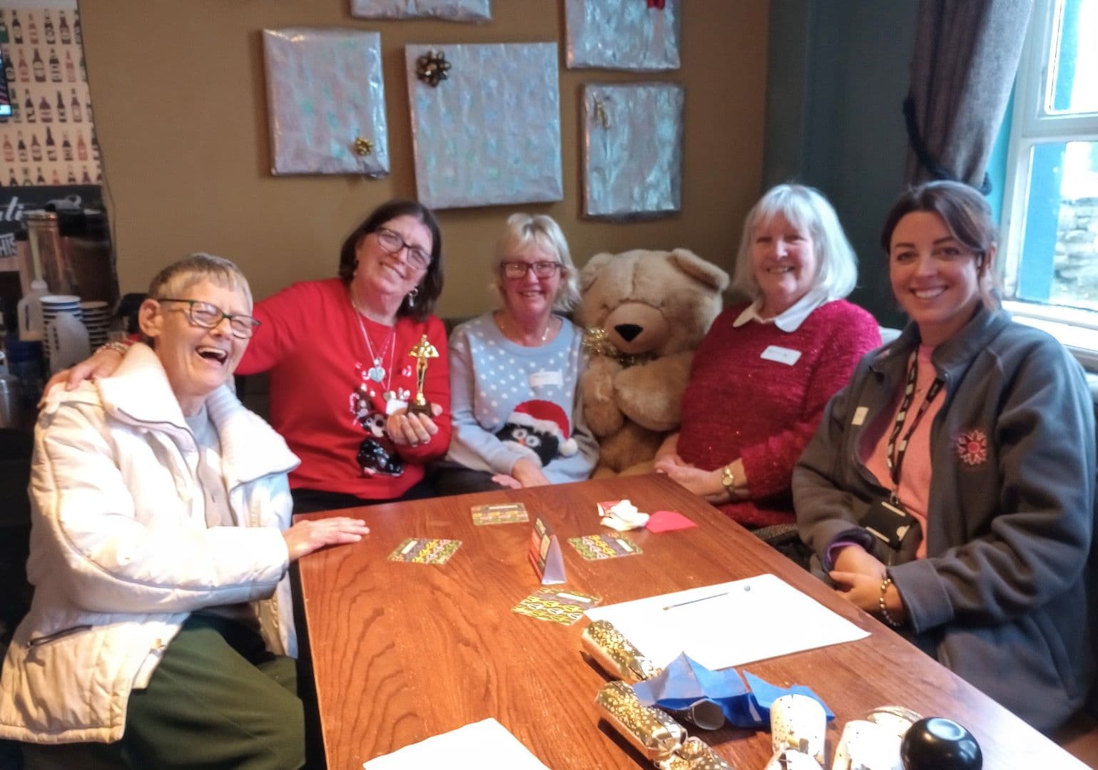 Five women smiling around a table with Christmas cards, a teddy bear, and festive decorations. - Home Instead